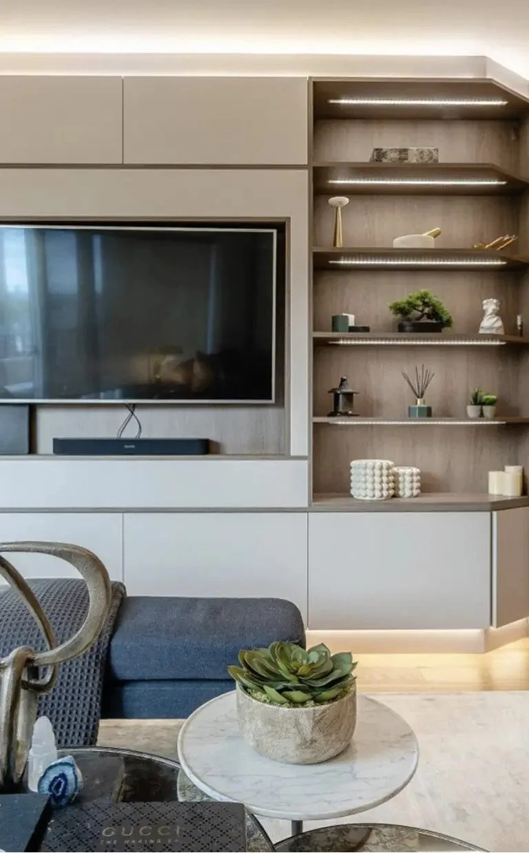 Living room with a large flat-screen TV mounted on light-colored cabinetry. Open shelving with decorative items, plants, books, and candles. A round marble coffee table with a succulent plant in a stone pot in the foreground. Part of a gray upholstered sofa and a woven chair are also visible.