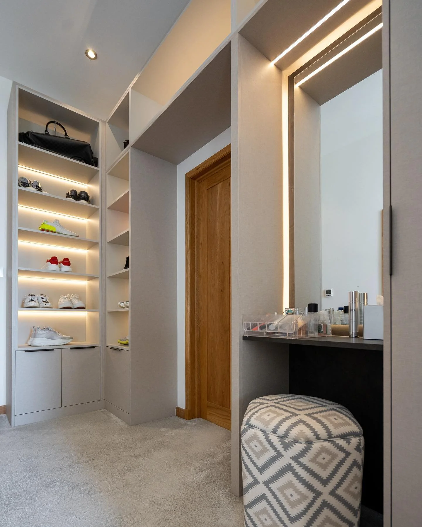 Modern walk-in closet with built-in shelves for shoes and accessories, a wooden door, a vanity with a mirror, and an ottoman with a geometric pattern.