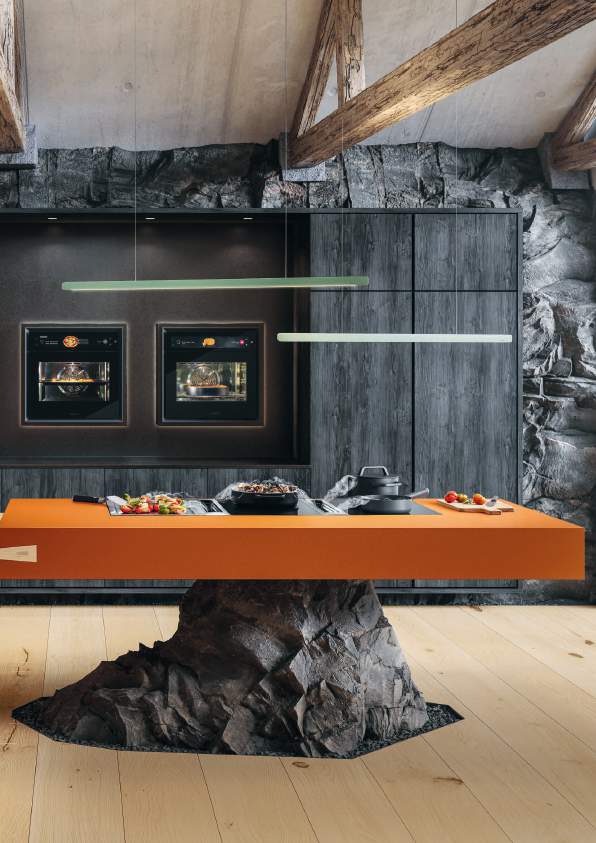 Modern kitchen with a large orange countertop supported by a rustic rock base, black stone wall, wood beams, and built-in ovens.