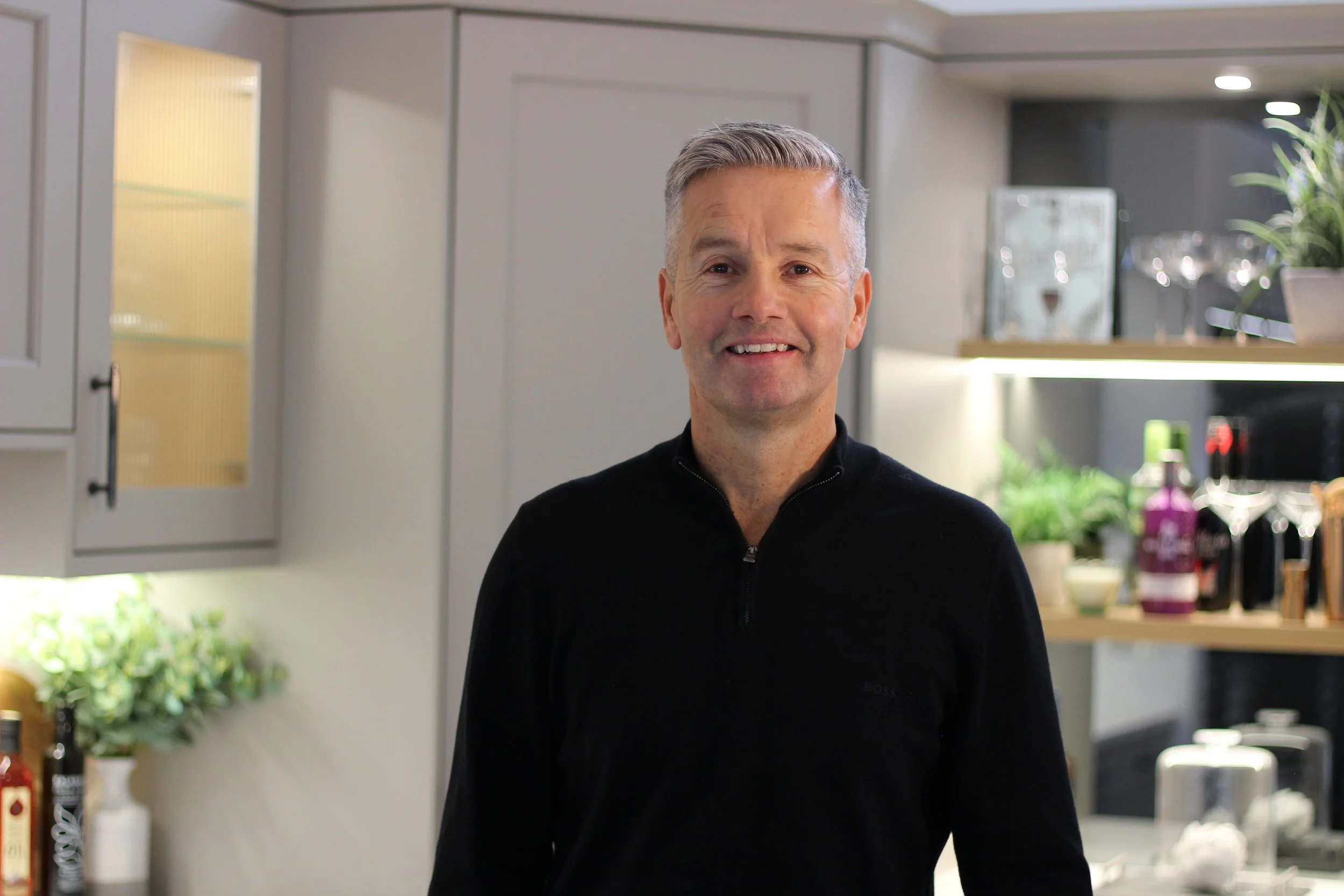 A smiling middle-aged man with gray hair standing in a modern kitchen with gray cabinets and shelves filled with bottles, plants, and glassware.