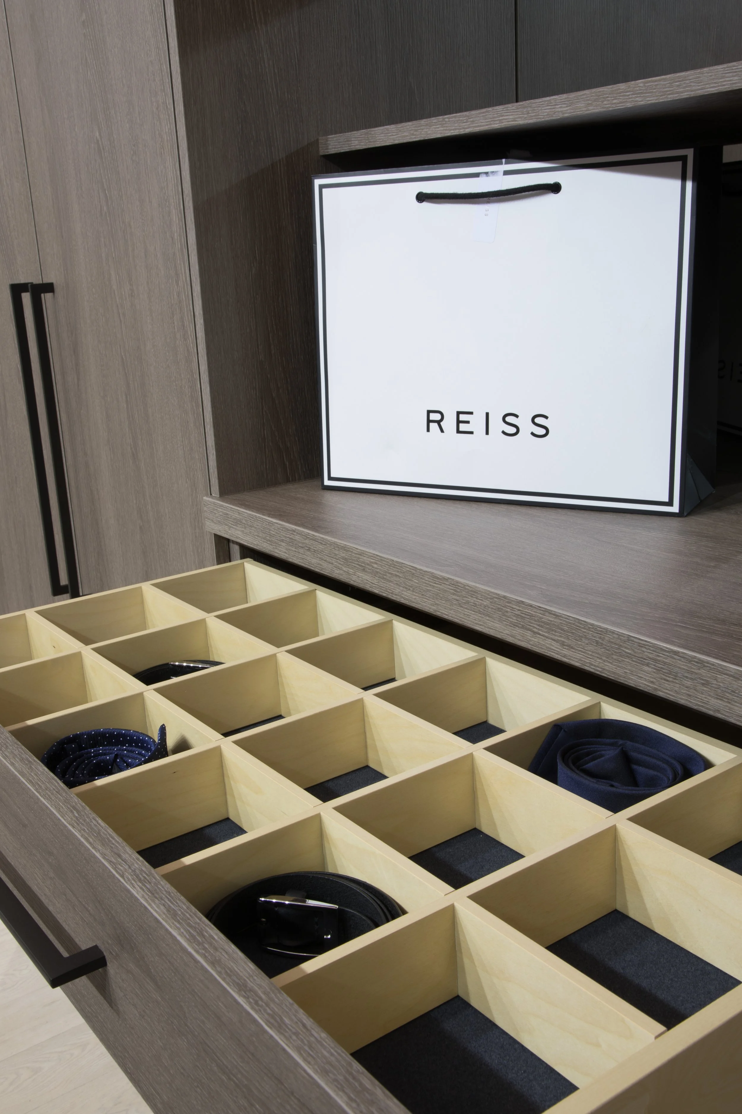 A drawer with compartments holding ties and belts, with a REISS shopping bag on the shelf above.