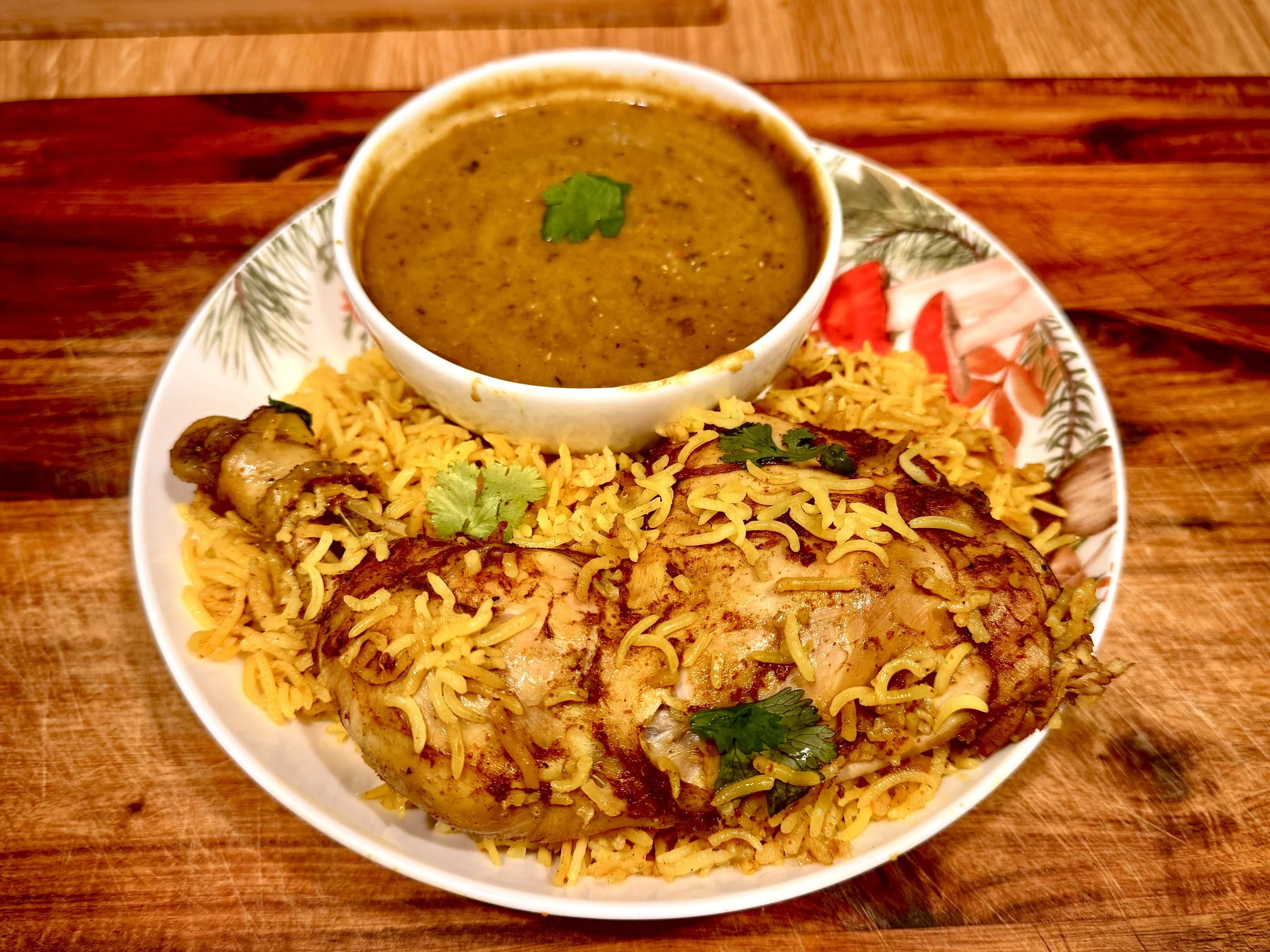Plate of yellow seasoned rice with chicken drumstick, grilled meat roll, and a bowl of brown curry garnished with cilantro, on a decorative plate with a floral and pinebranch pattern.