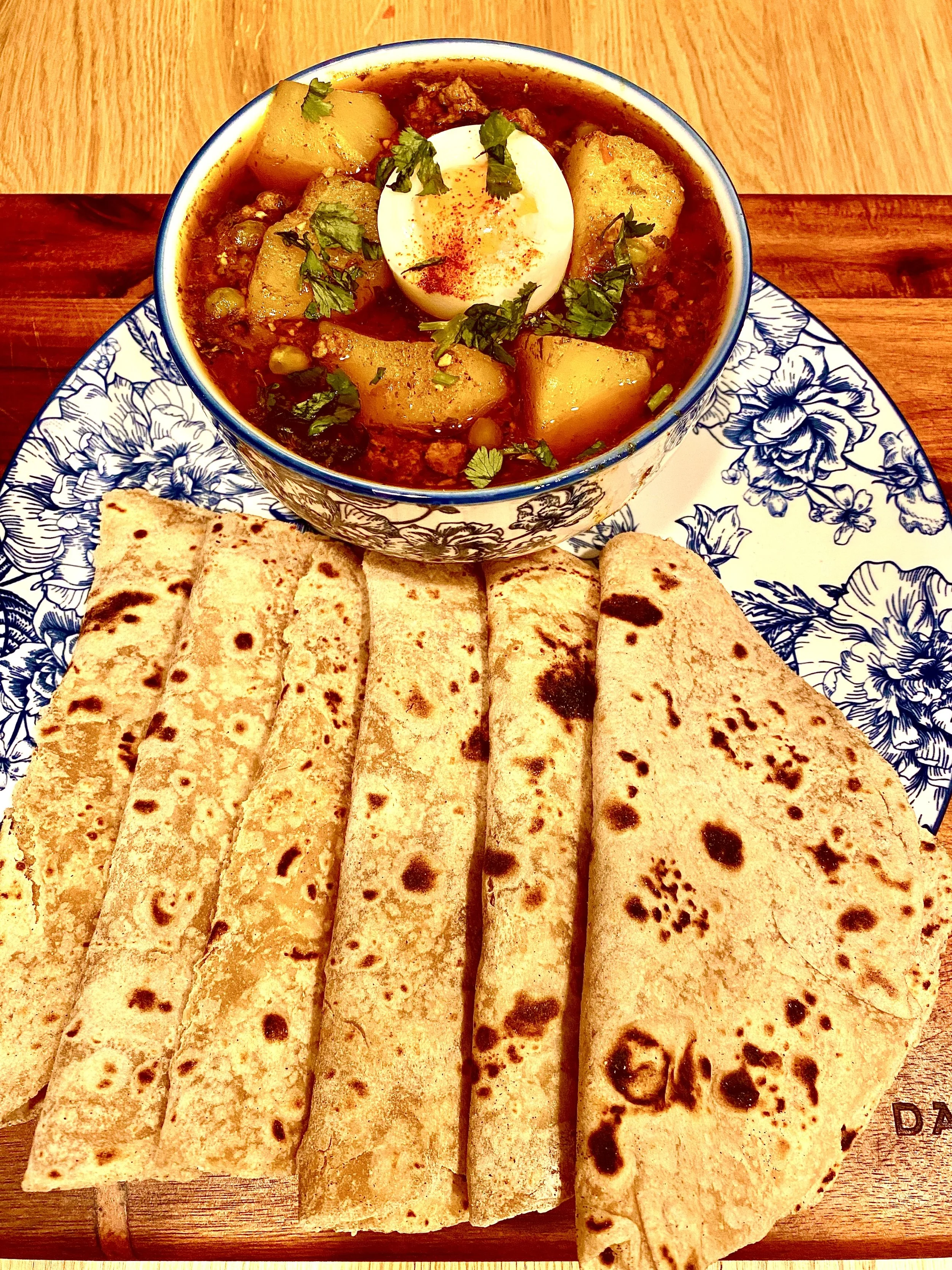 Indian meal consisting of naan bread and a bowl of vegetable curry with potatoes, chickpeas, topped with a boiled egg and garnished with cilantro on a decorative plate.