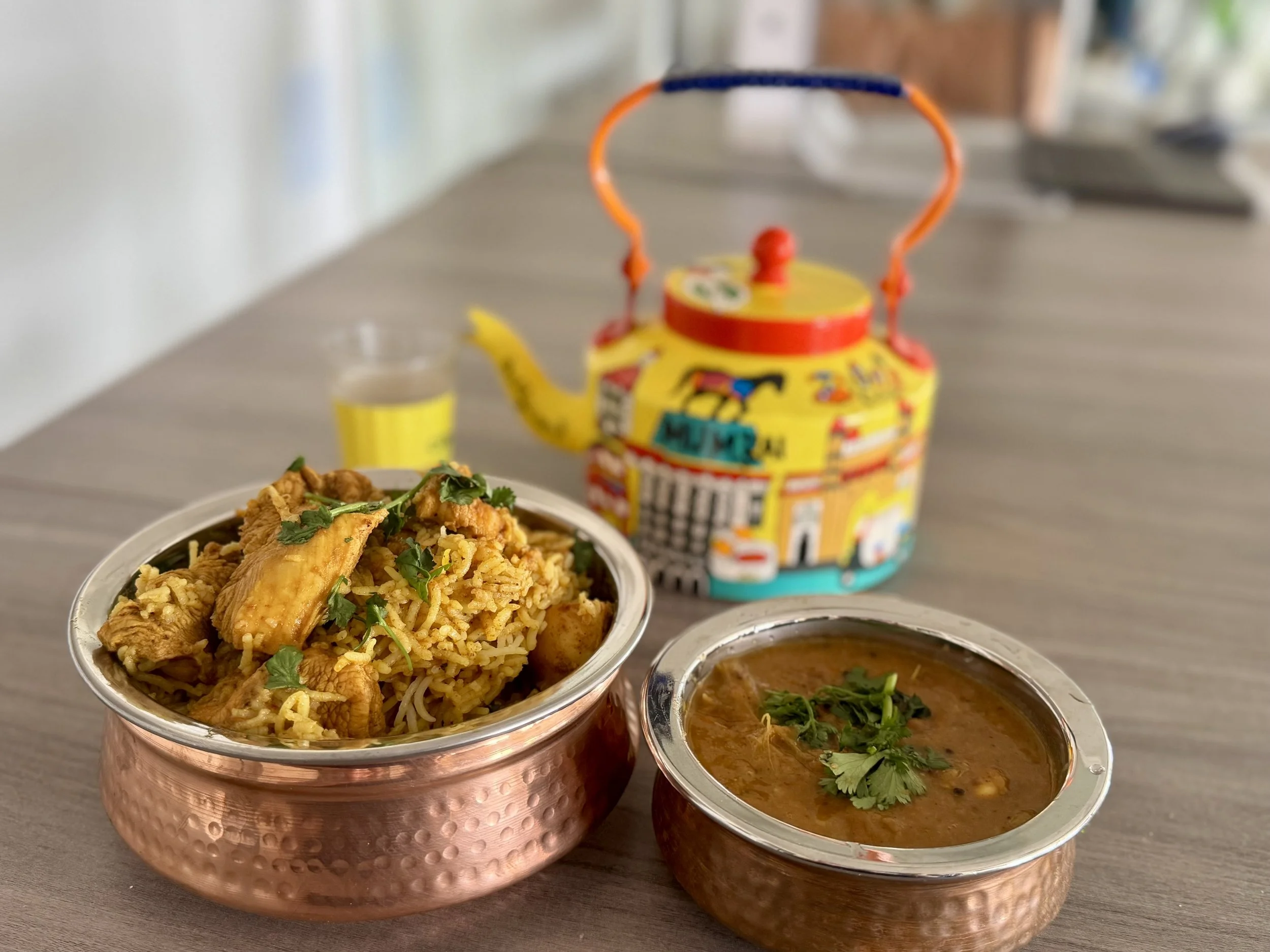 Copper bowls containing rice and chicken dish garnished with cilantro, and spicy curry garnished with cilantro, on a wooden table with a colorful teapot and a glass of water in the background.
