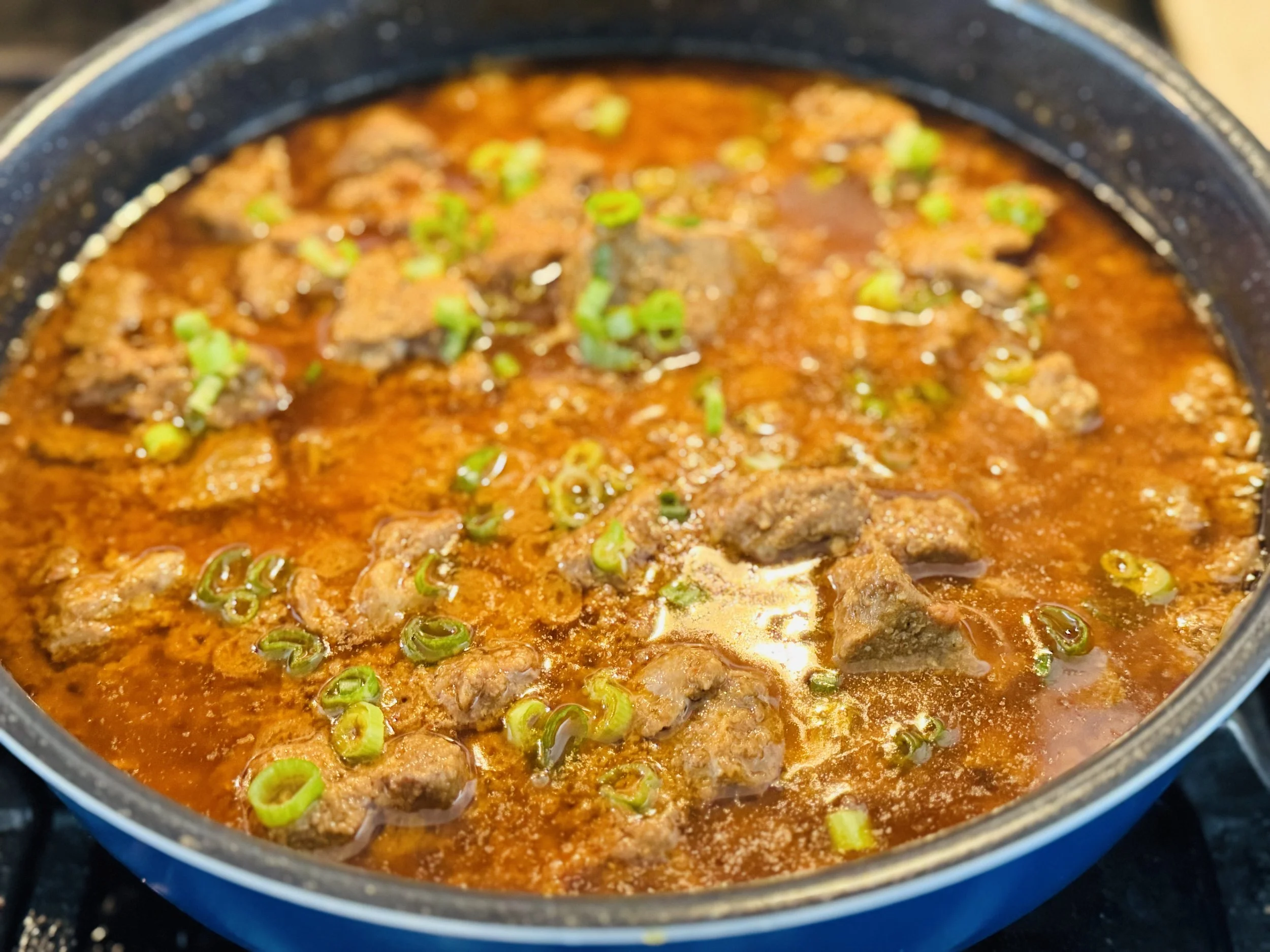 A pot of beef stew with chunks of meat and green onions in a red broth.
