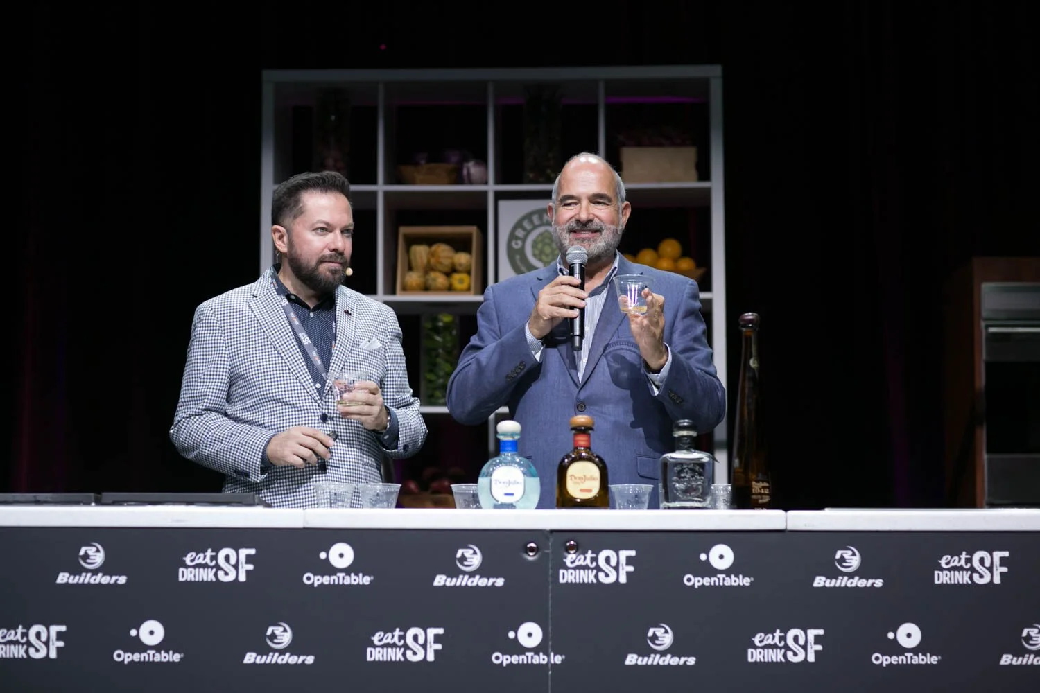 Two men standing behind a table, one holding a glass and speaking into a microphone, with bottles of liquor and glasses on the table, at a promotional event for spirits.