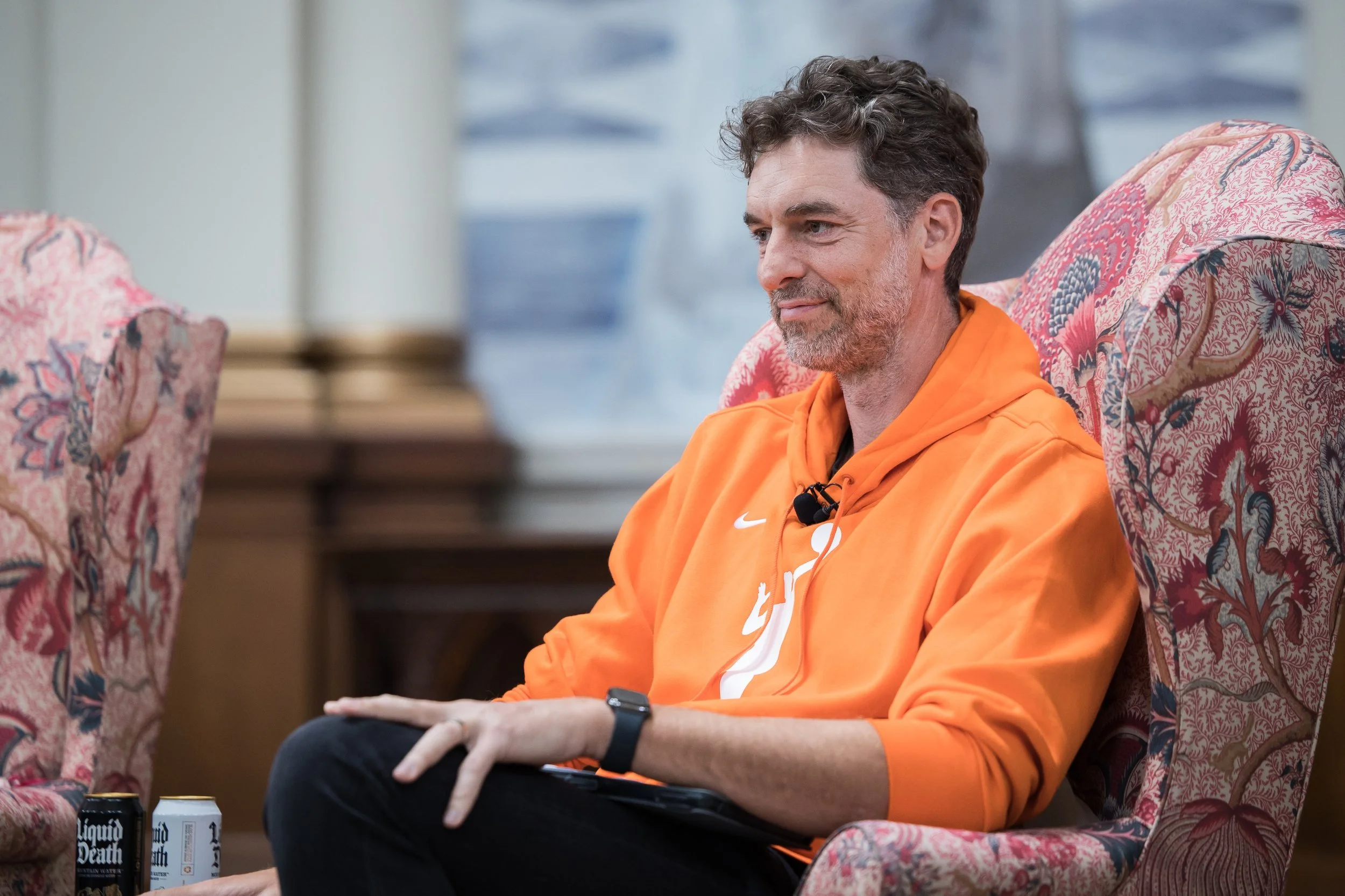 A man with curly hair and a beard, wearing an orange hoodie, sitting on a patterned armchair.