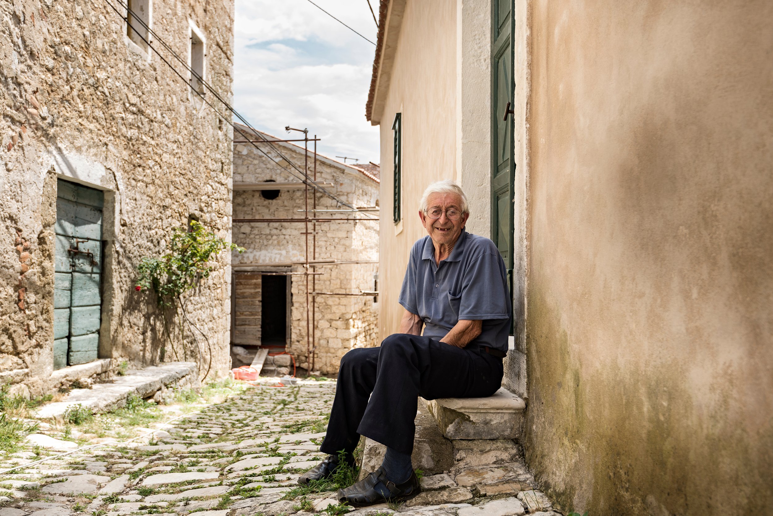 An elderly man with glasses, white hair, and a blue shirt sitting on a stone step outside in a narrow alley with old stone and plaster buildings.