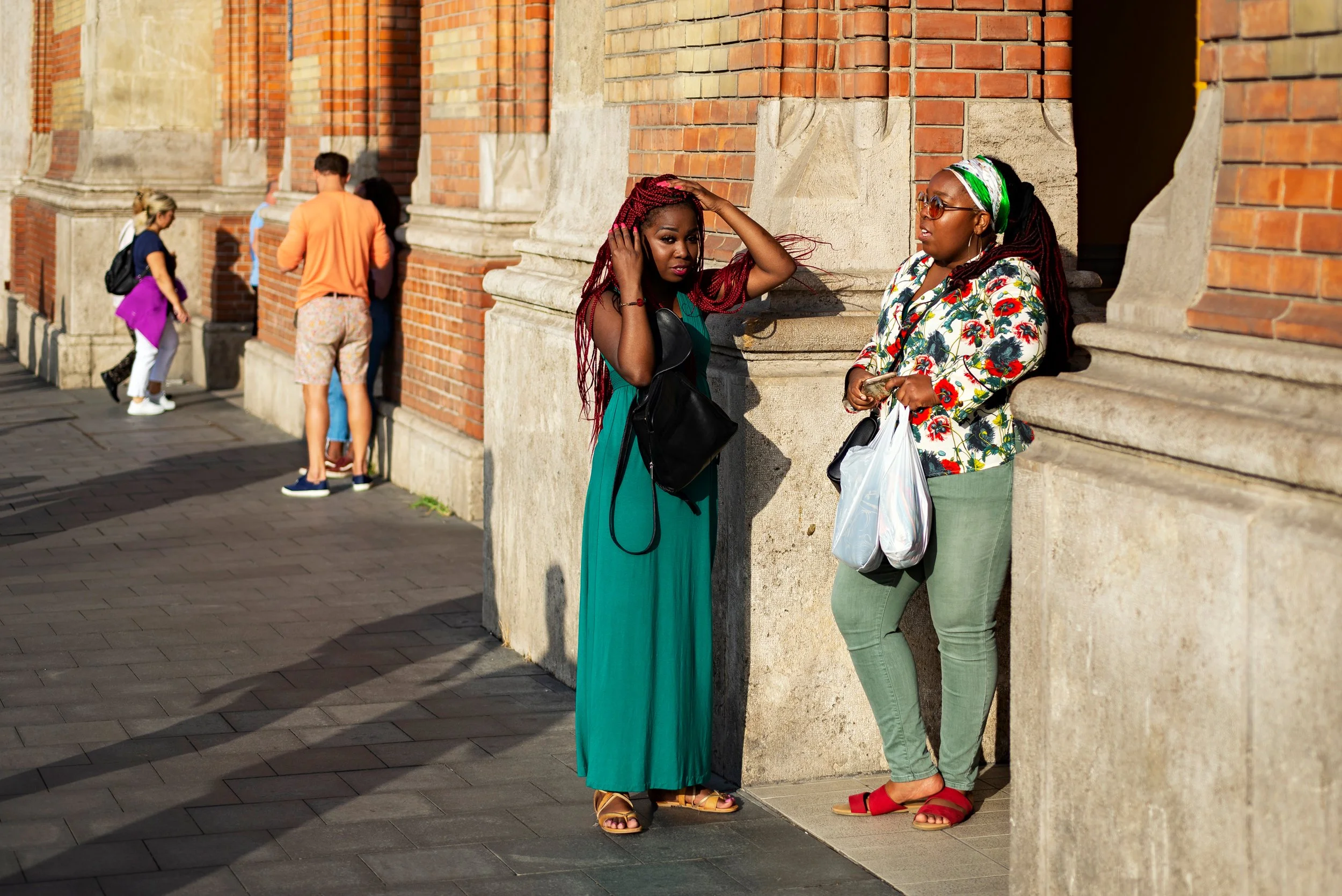 Two women stand against a brick wall in sunlight, one with red braided hair wearing a teal dress, the other with sunglasses and a floral top, holding a plastic bag. Three people stand in line nearby.