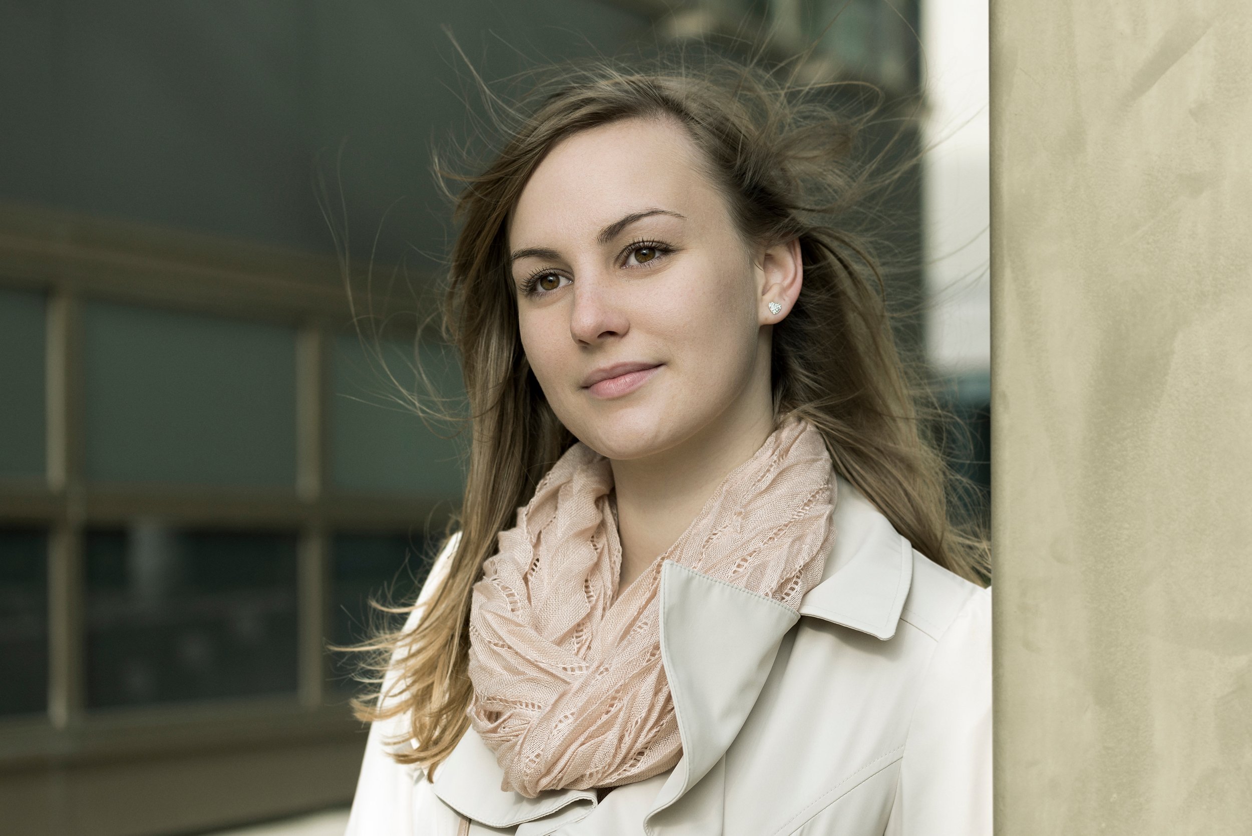 A young woman with brown hair, light makeup, and hoop earrings standing outdoors near a yellow architectural pillar with a modern building in the background, leaning against the pillar and smiling softly.