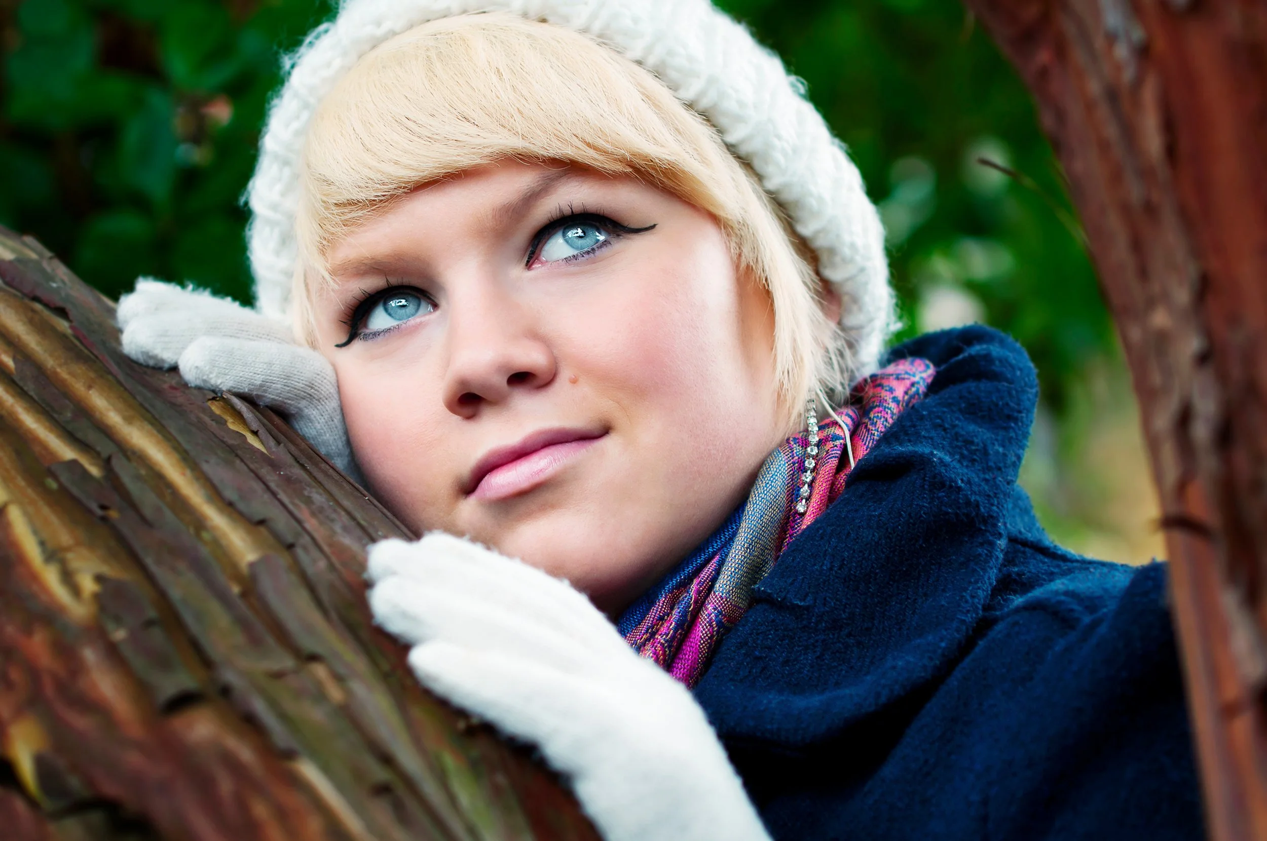 A young woman with blonde hair and blue eyes, wearing a white knit hat and gloves, resting her head on a tree branch outdoors surrounded by green foliage.