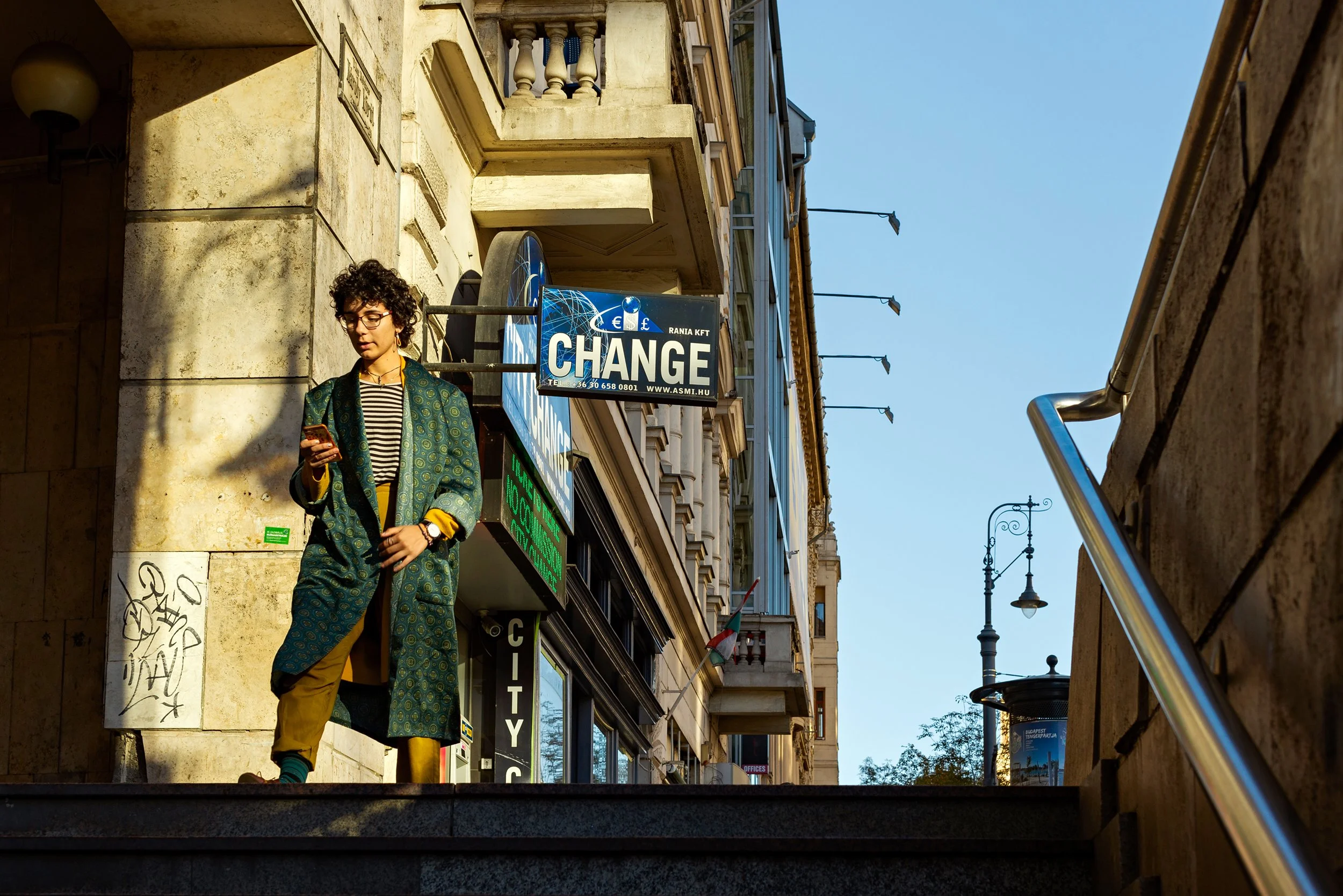 A young person with curly hair and glasses standing under a building overhang, looking at their phone. They are wearing a patterned coat, striped shirt, and yellow trousers. There is a sign that reads 'CHANGE' and other signs on the building.