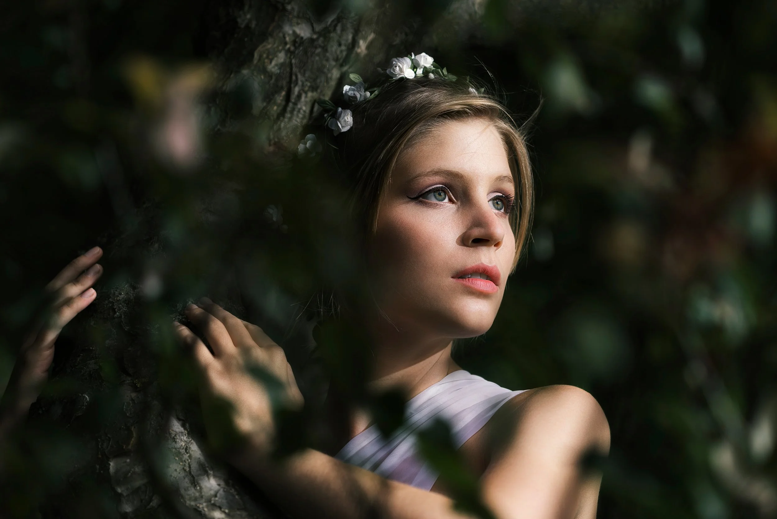 A young woman with blonde hair and blue eyes wearing a white top and a flower crown, resting her head against a tree outdoors with greenery surrounding her.