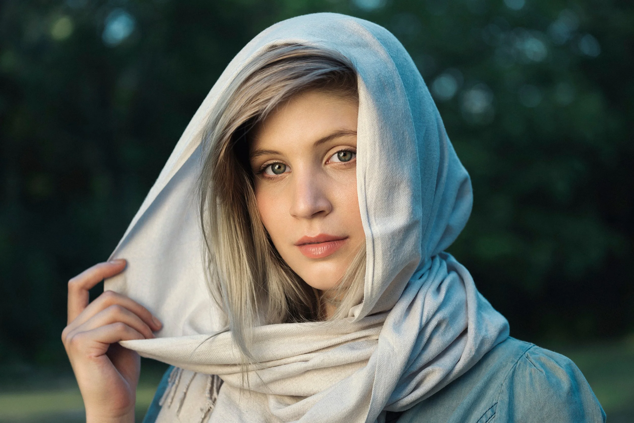 Young woman with blonde hair and blue eyes wearing a gray hoodie and a light-colored scarf, outdoors with blurred greenery background.