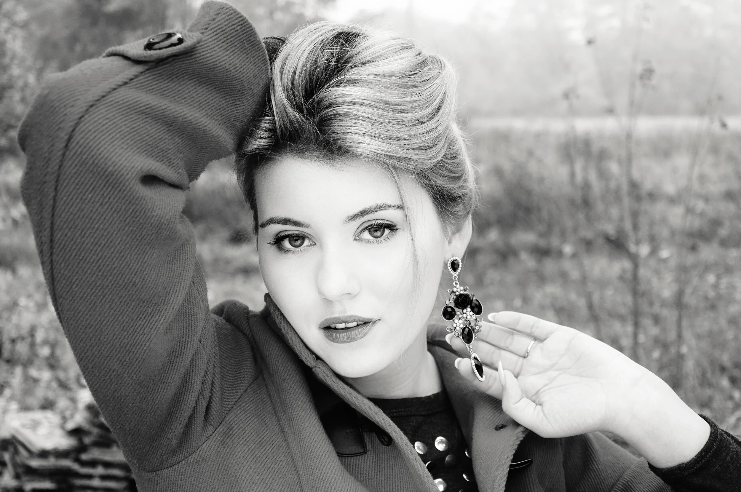 A woman with short hair, wearing earrings and jewelry, holding a large gemstone necklace, outdoors with blurred trees in the background.