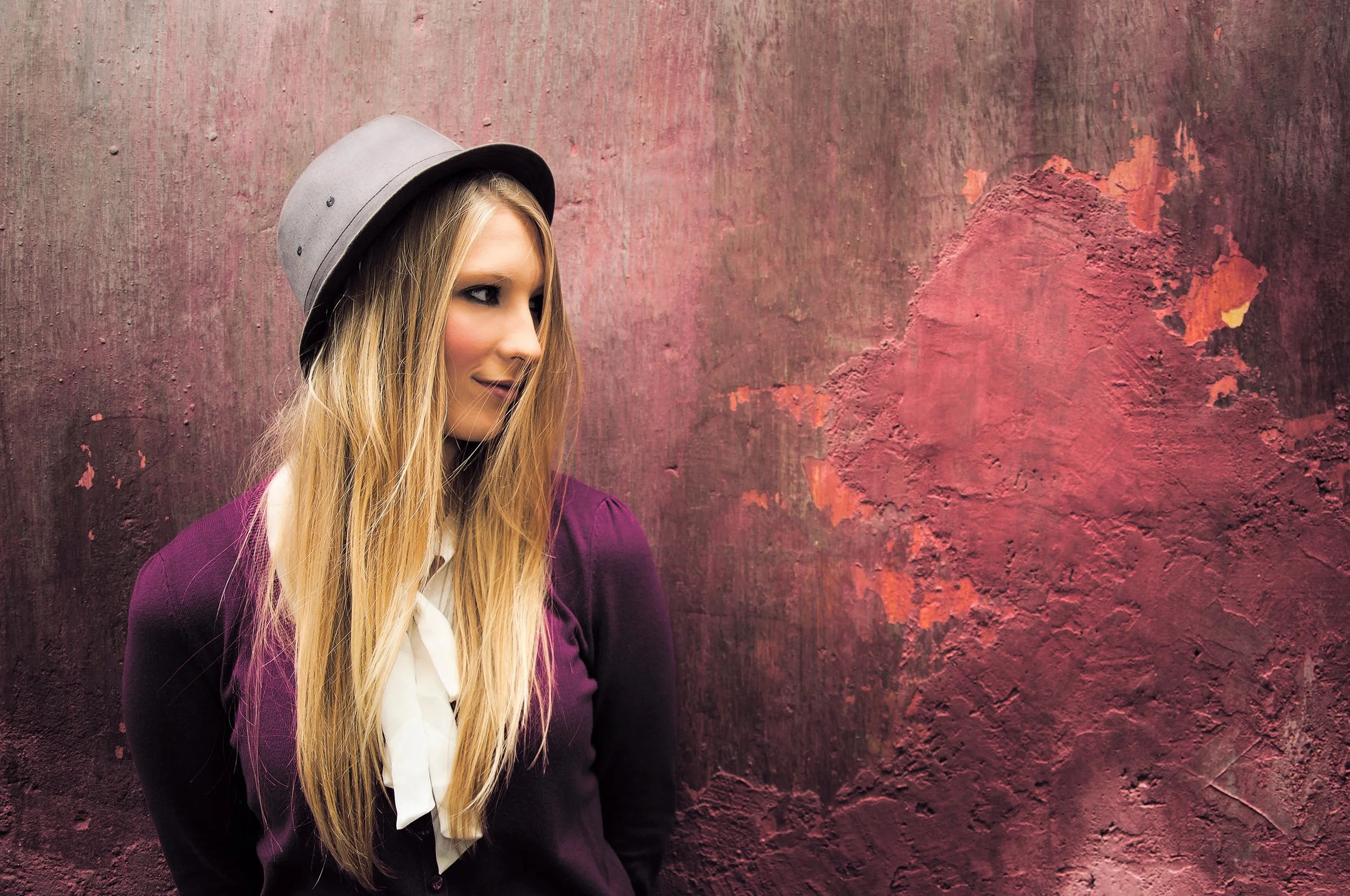 A woman with long blonde hair wearing a gray hat, a white blouse, and a dark purple blazer, standing against a textured pinkish-red wall.