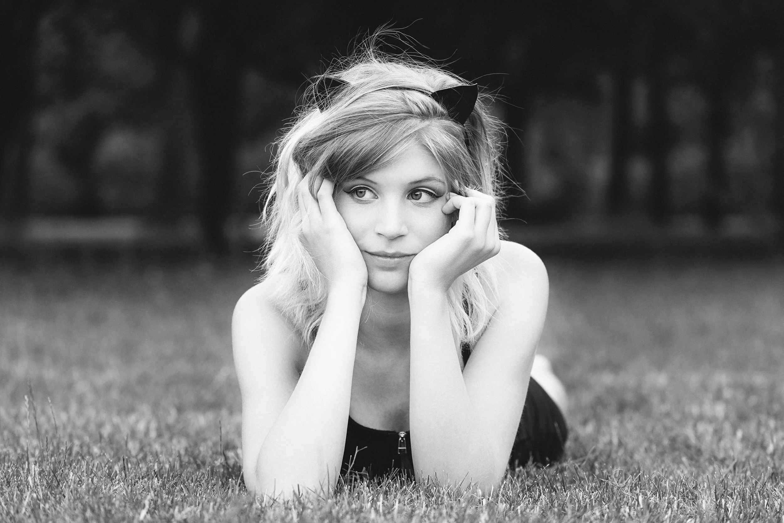 Black and white photo of a young woman with light hair, wearing a hairband with cat ears, lying on her stomach on grass in a park, resting her face in her hands and looking to the side.