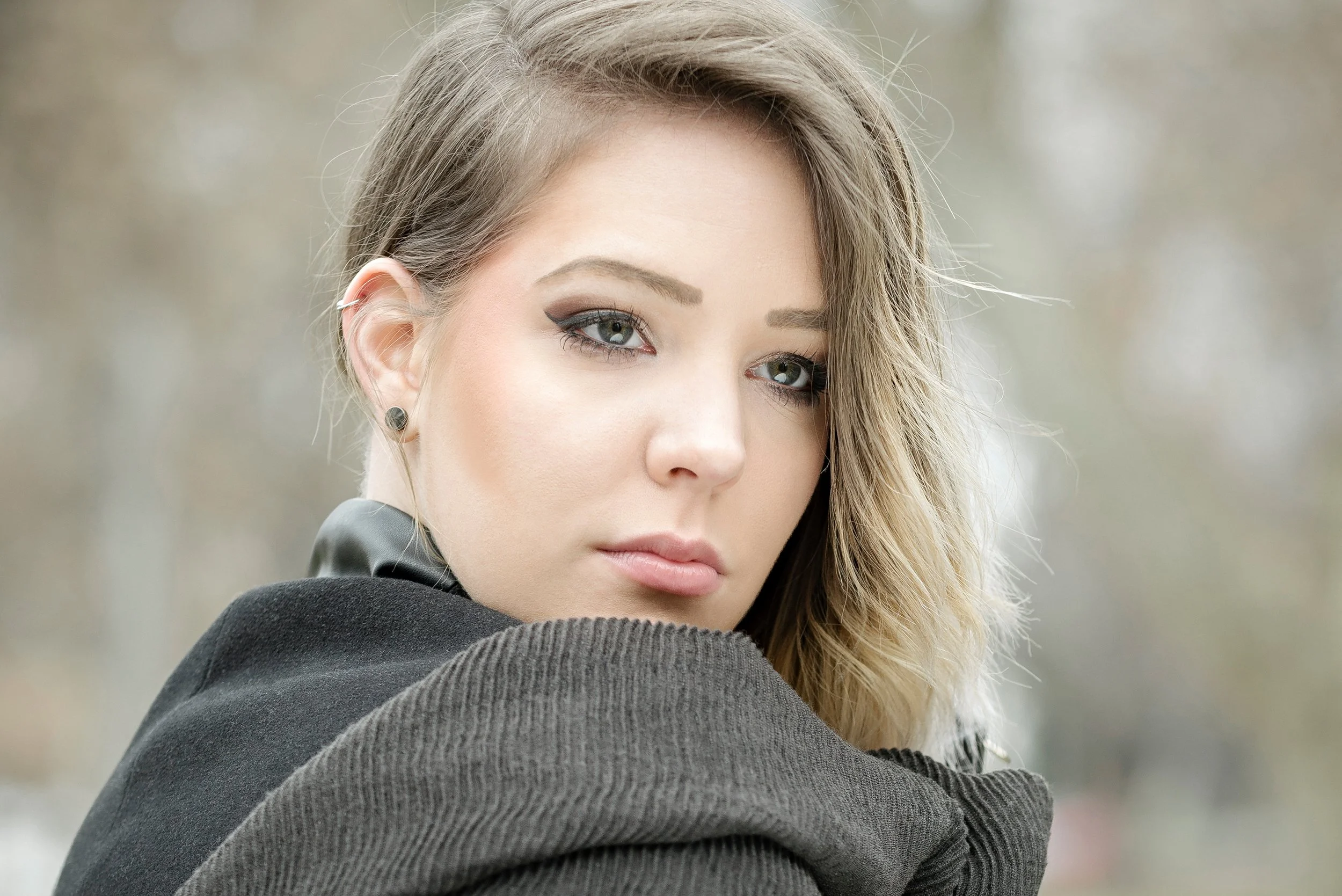 A young woman with blonde hair, blue eyes, and makeup looking directly at the camera with a neutral expression, wearing a black jacket and a striped shirt.