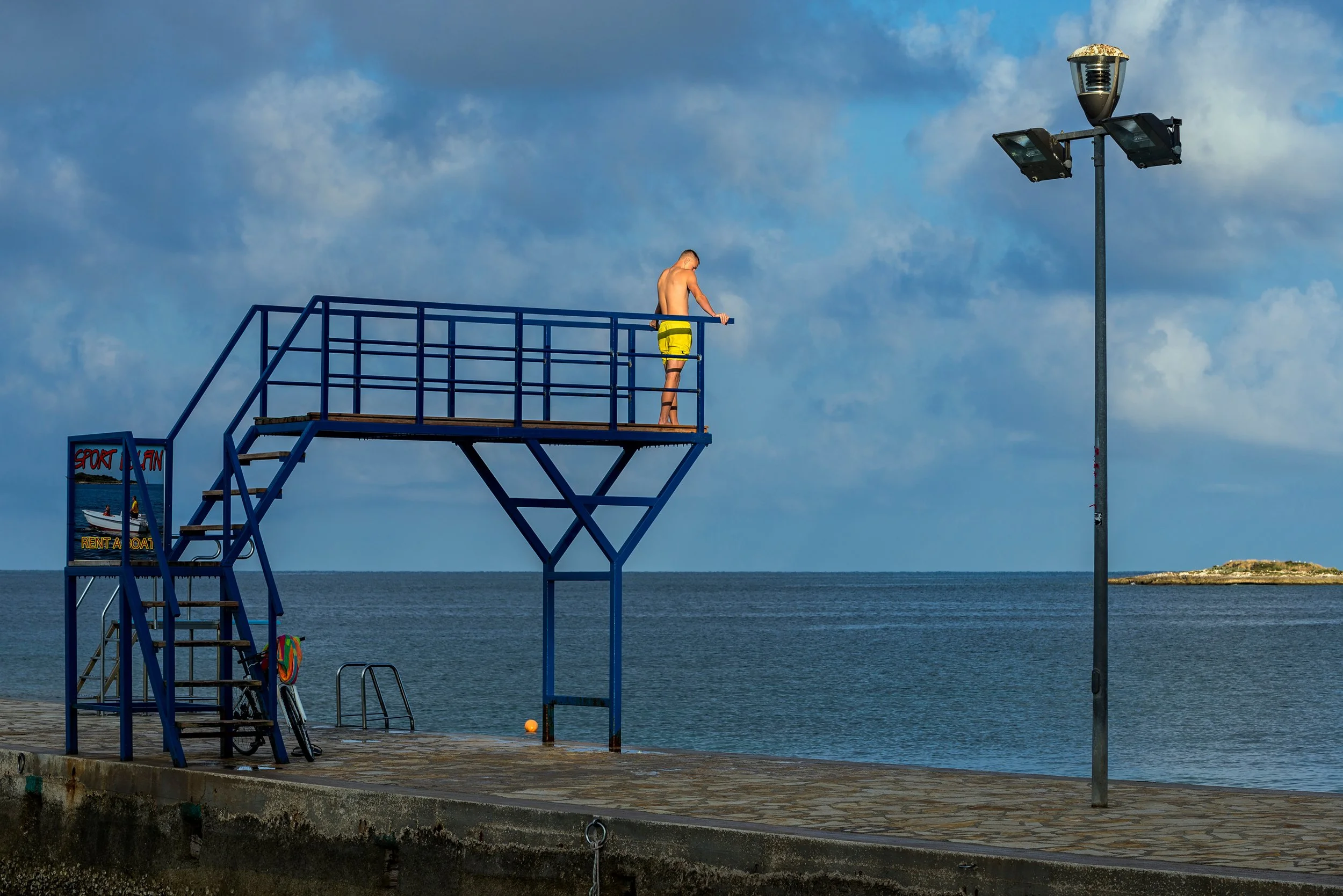 A person standing on a high diving platform at the beach, with the ocean and a small island in the background, and a street lamp nearby.