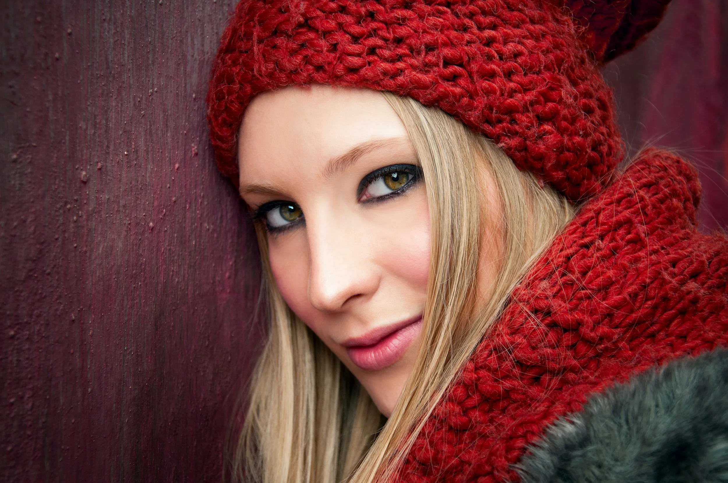 A woman with blonde hair, green eyes, and makeup, wearing a large red knit hat and a matching red knit scarf, leaning against a textured red wooden wall.