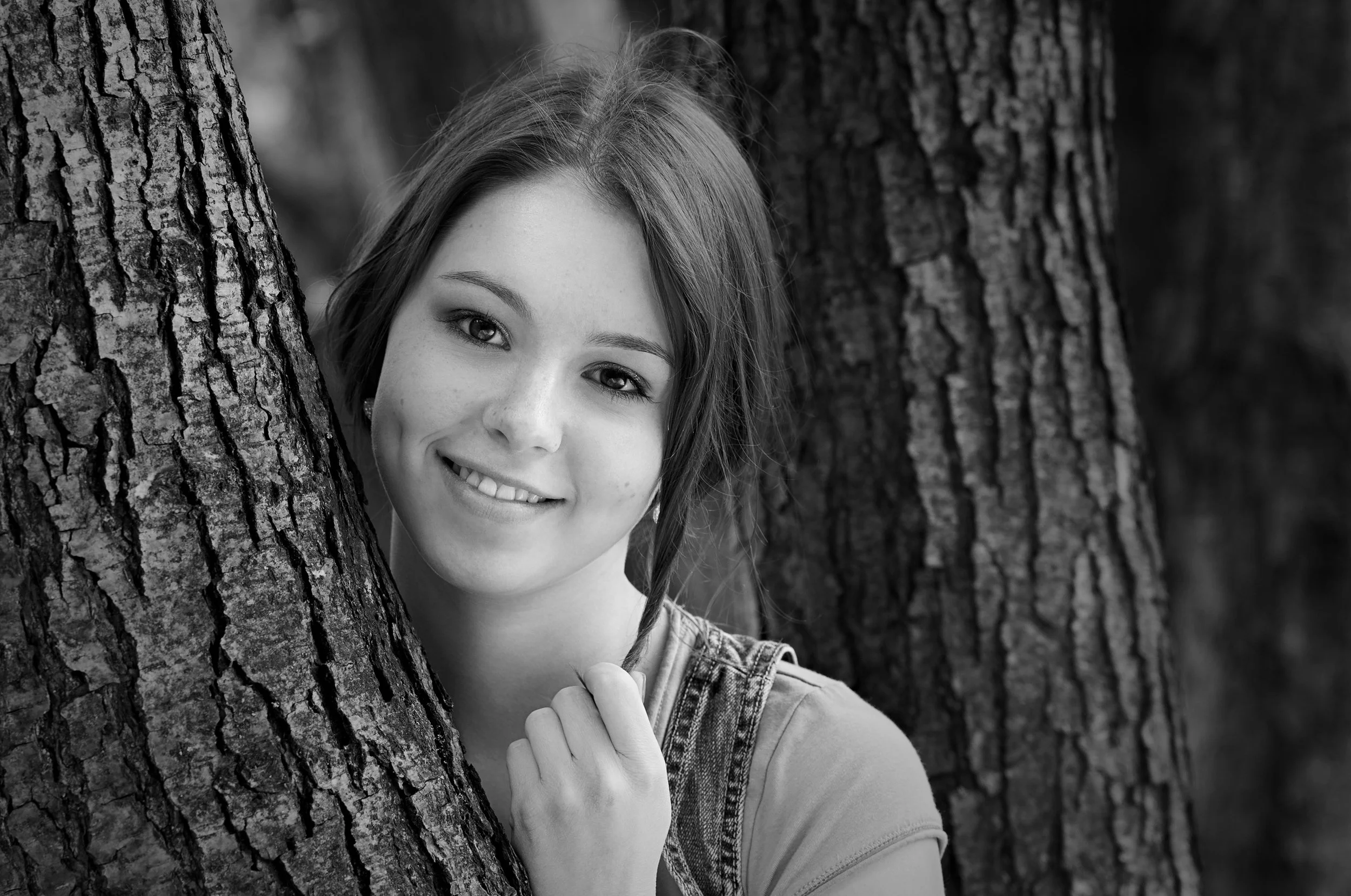 A black and white photo of a woman with light skin and long hair, smiling, hiding behind a tree, with her face partially visible.