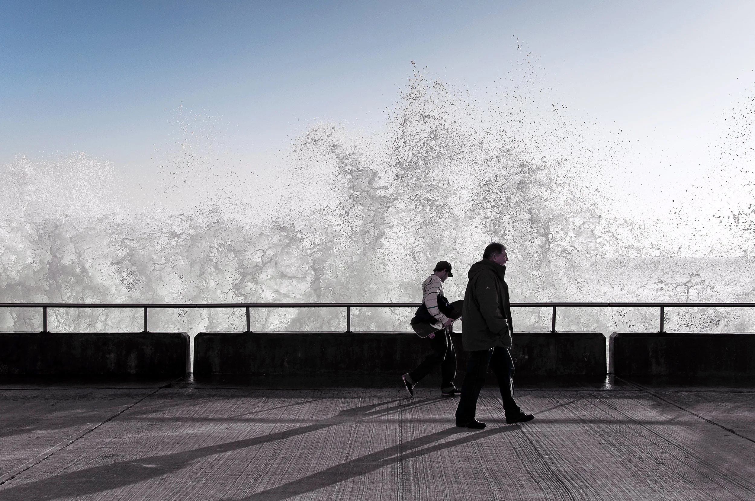 People walking along a promenade by an ocean with large waves crashing against the rocks, under a clear sky.