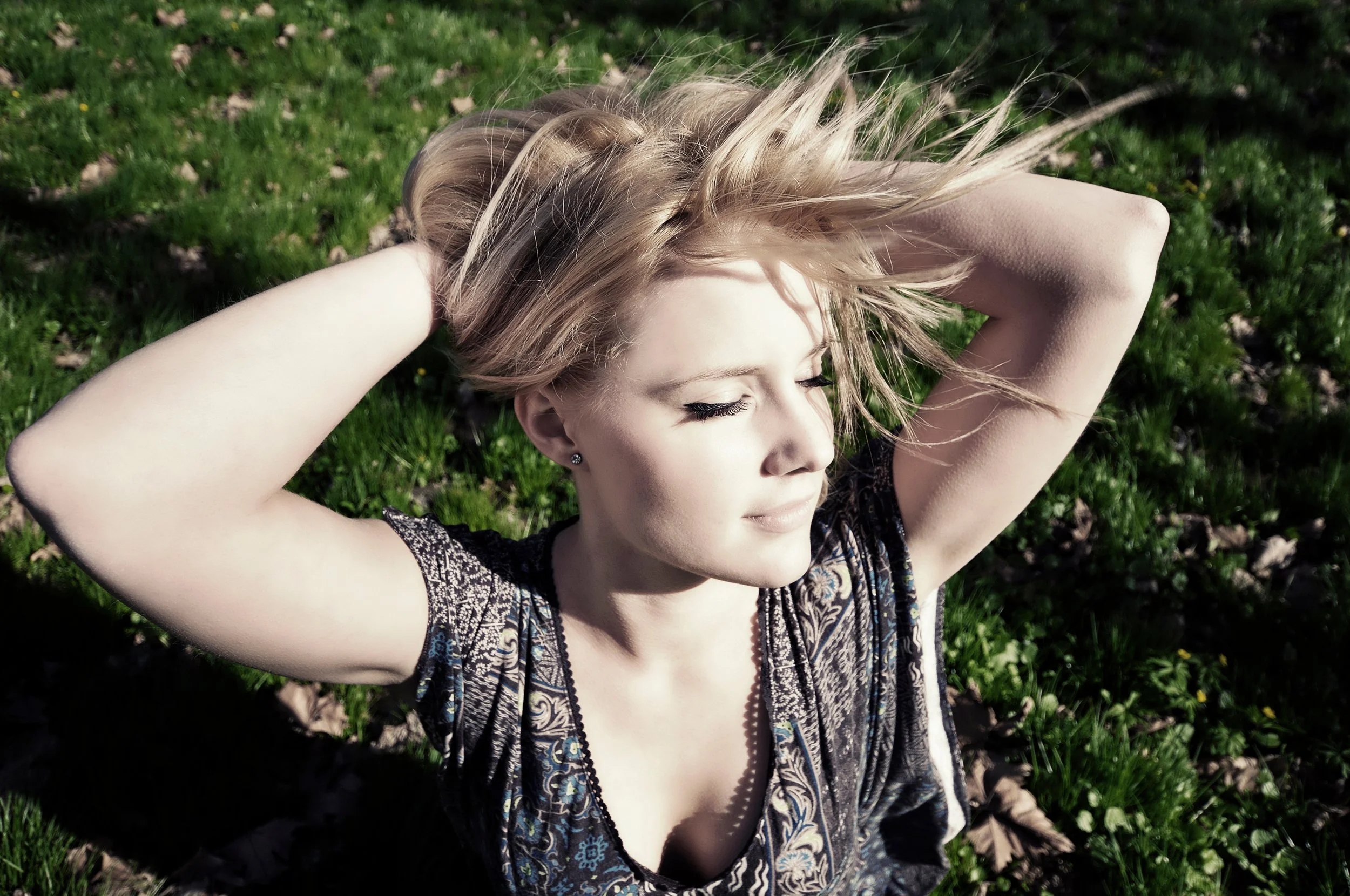A woman with blonde hair lying on grass with her eyes closed, arms behind her head, in sunlight.