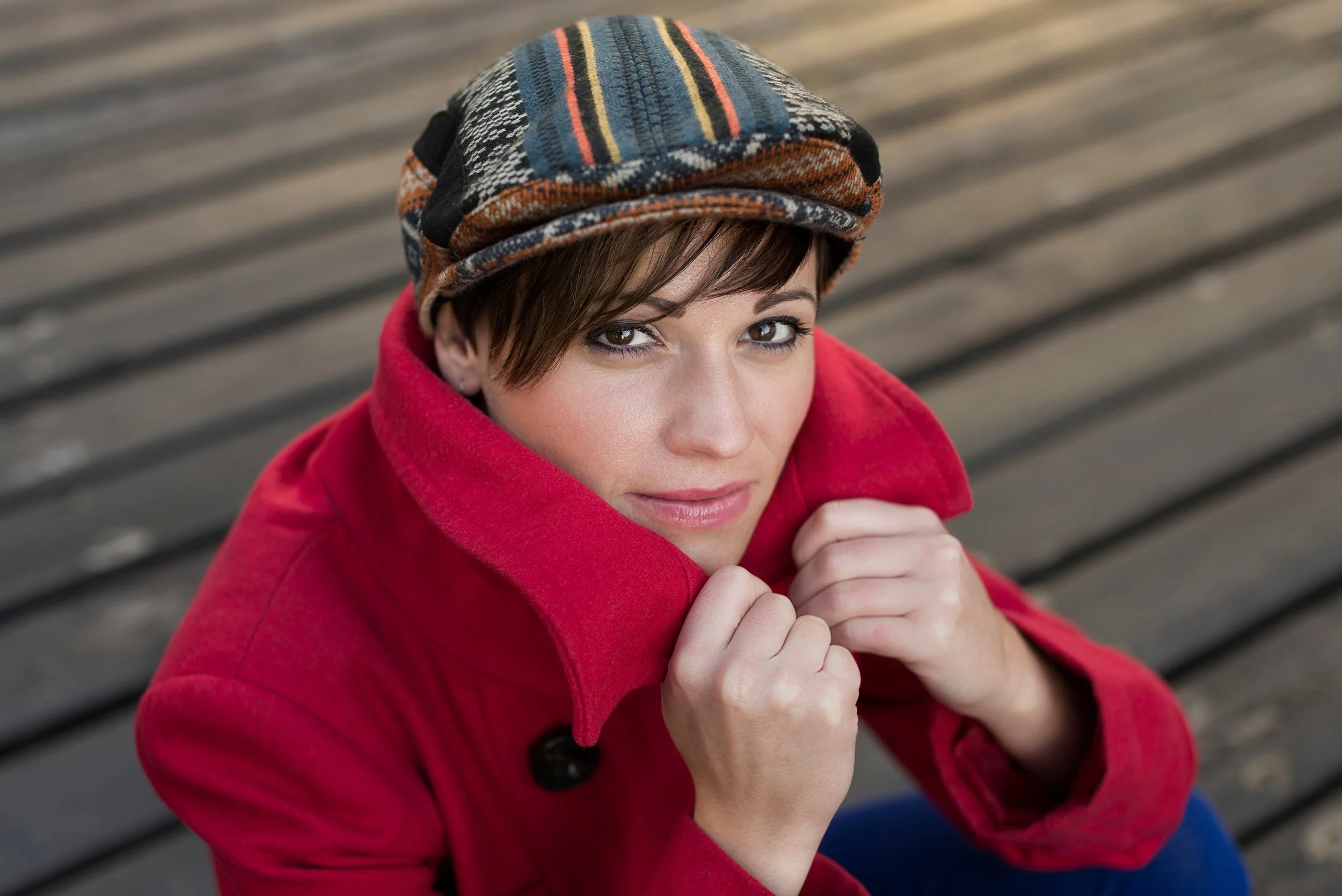 A woman with short brown hair and brown eyes, wearing a colorful striped knit hat and a red coat, sitting on a bench outdoors.