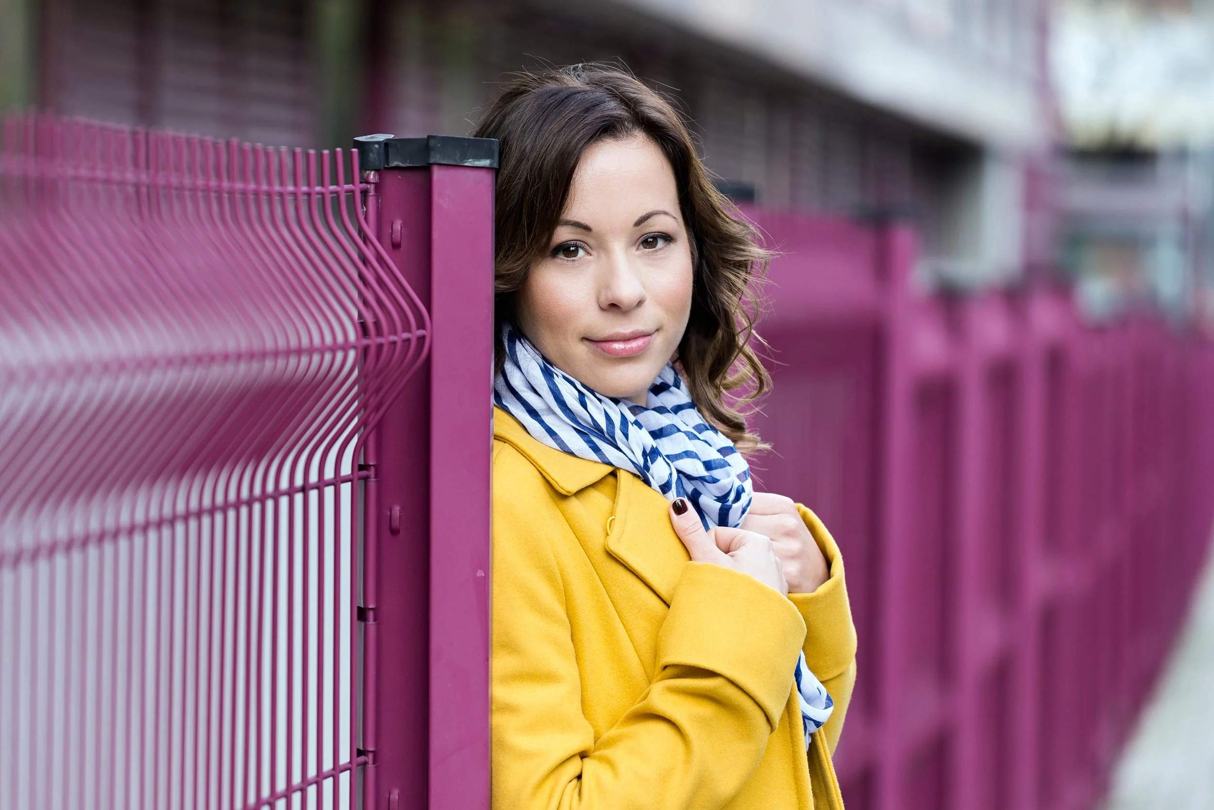A woman with brown hair, wearing a yellow coat and a blue-and-white striped scarf, leaning next to a pink metal fence.