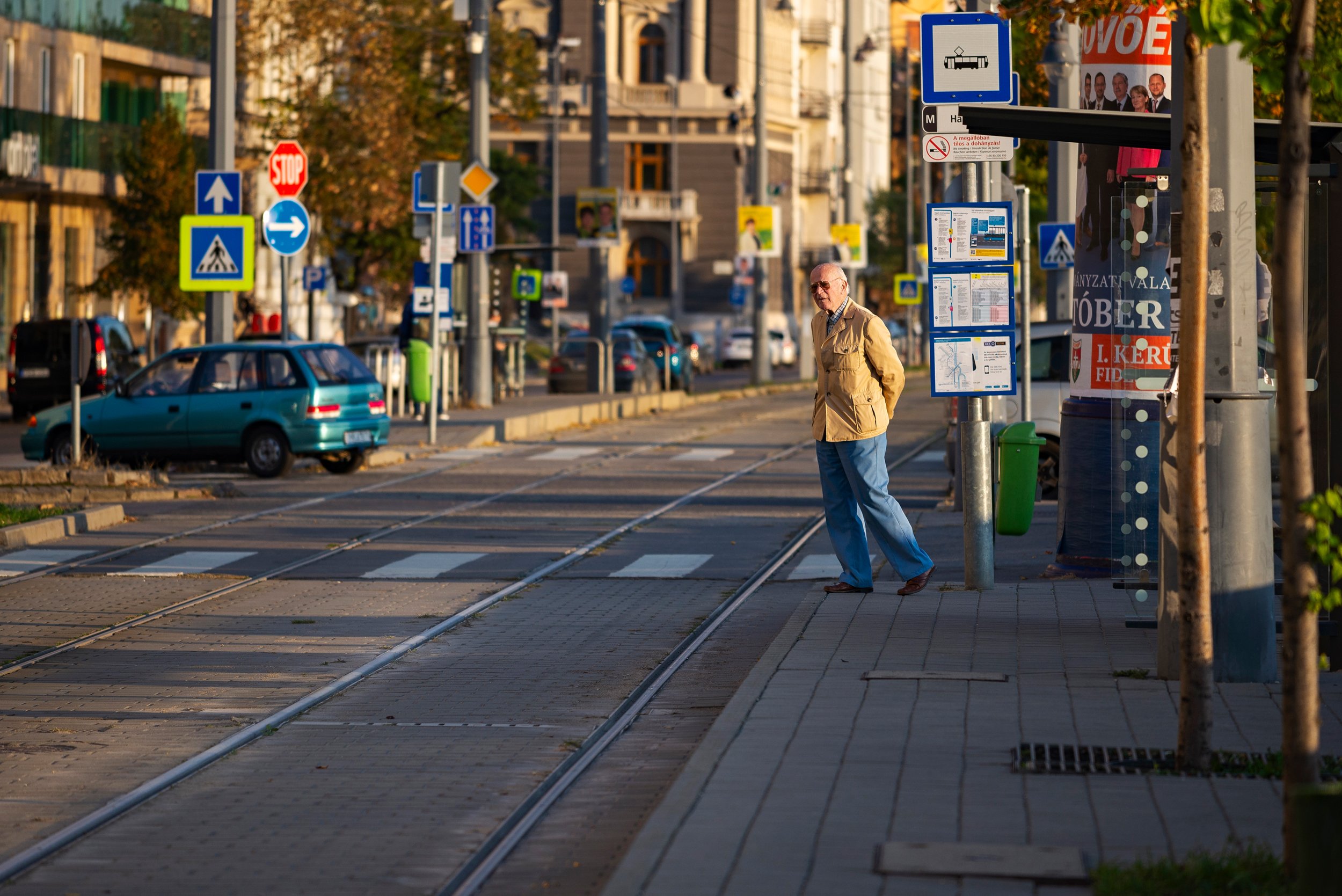 Older man in a beige jacket and blue pants waiting at a tram stop on a city street during the daytime.