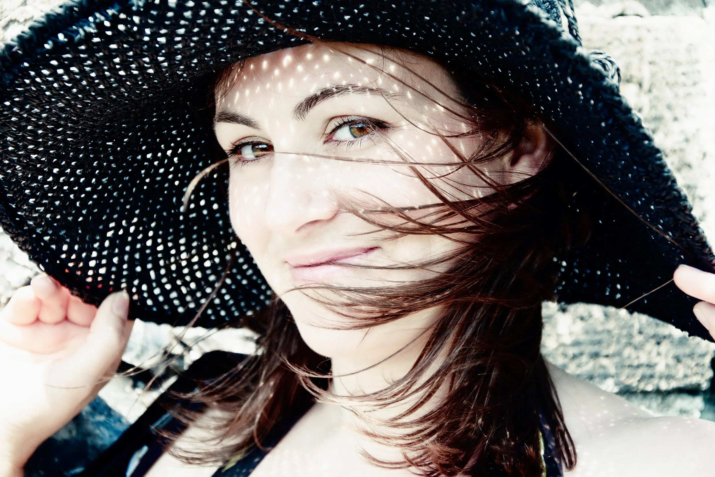 A woman with brown hair and brown eyes wearing a large black hat with white dots, outdoors in sunlight, with her hair blowing across her face.