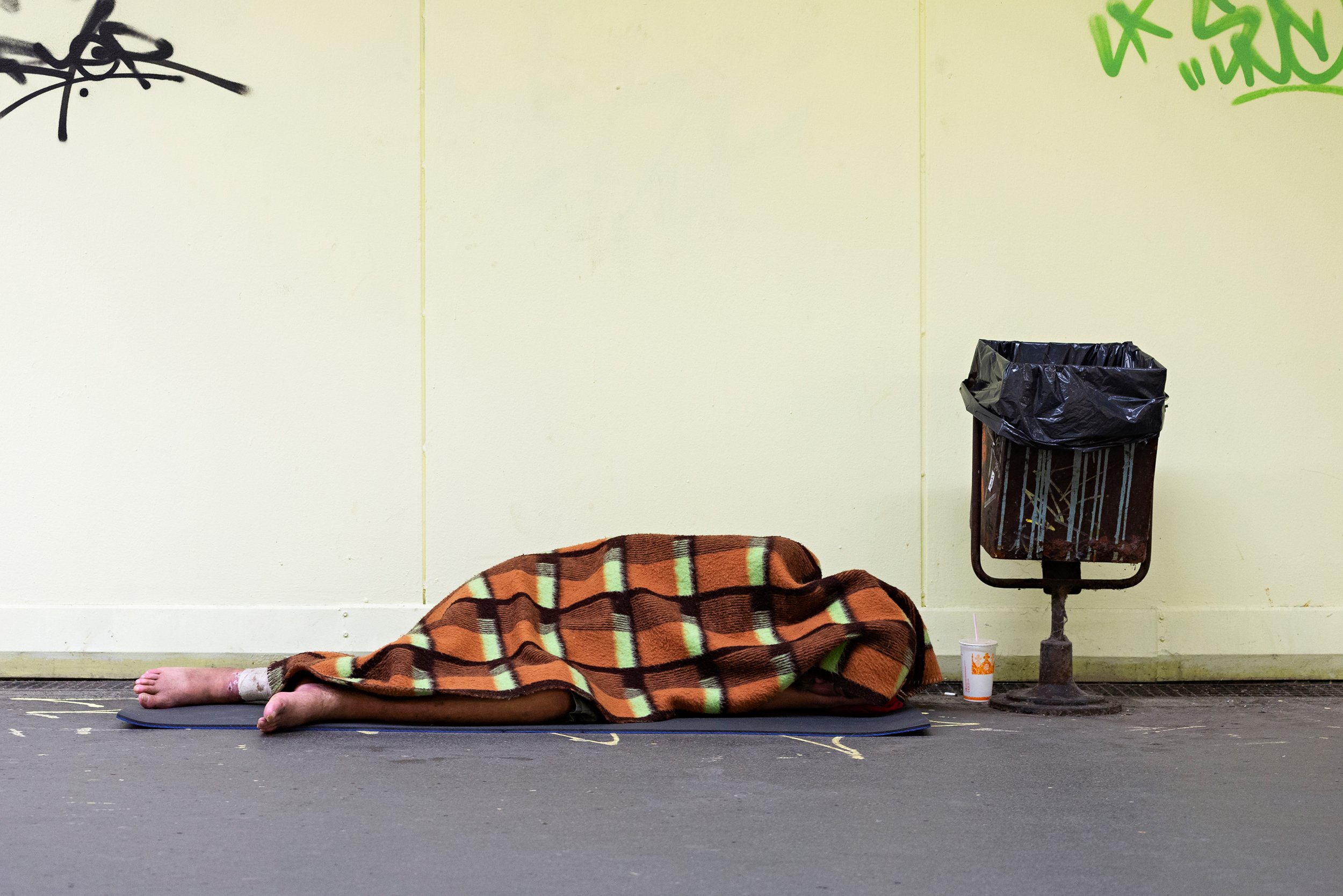 A person lying on the floor with only legs, feet, and toes visible, covered by a brown, orange, and white checkered blanket. There is a mat underneath them, and a small bandage is on their ankle. To the right is a trash can with a black plastic bag a