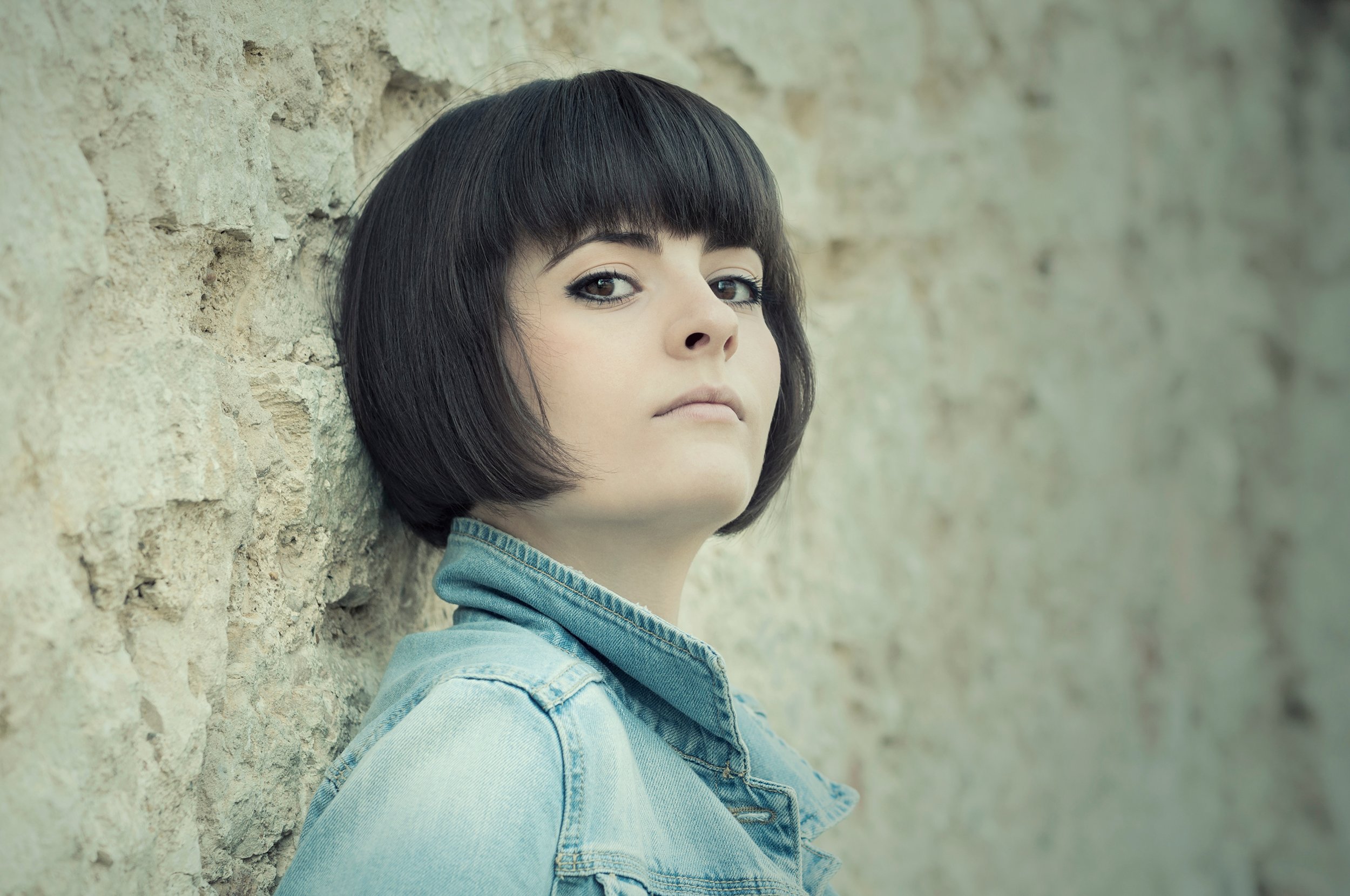 A young woman with short dark hair and a fringe, wearing a denim jacket, leaning against a textured stone wall with neutral expression.