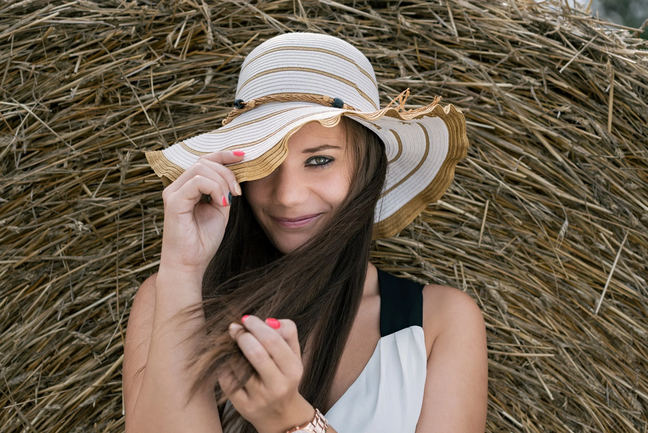 Woman with long dark hair lying on a large pile of hay, wearing a wide-brimmed straw hat, smiling, and holding the brim of her hat with one hand.