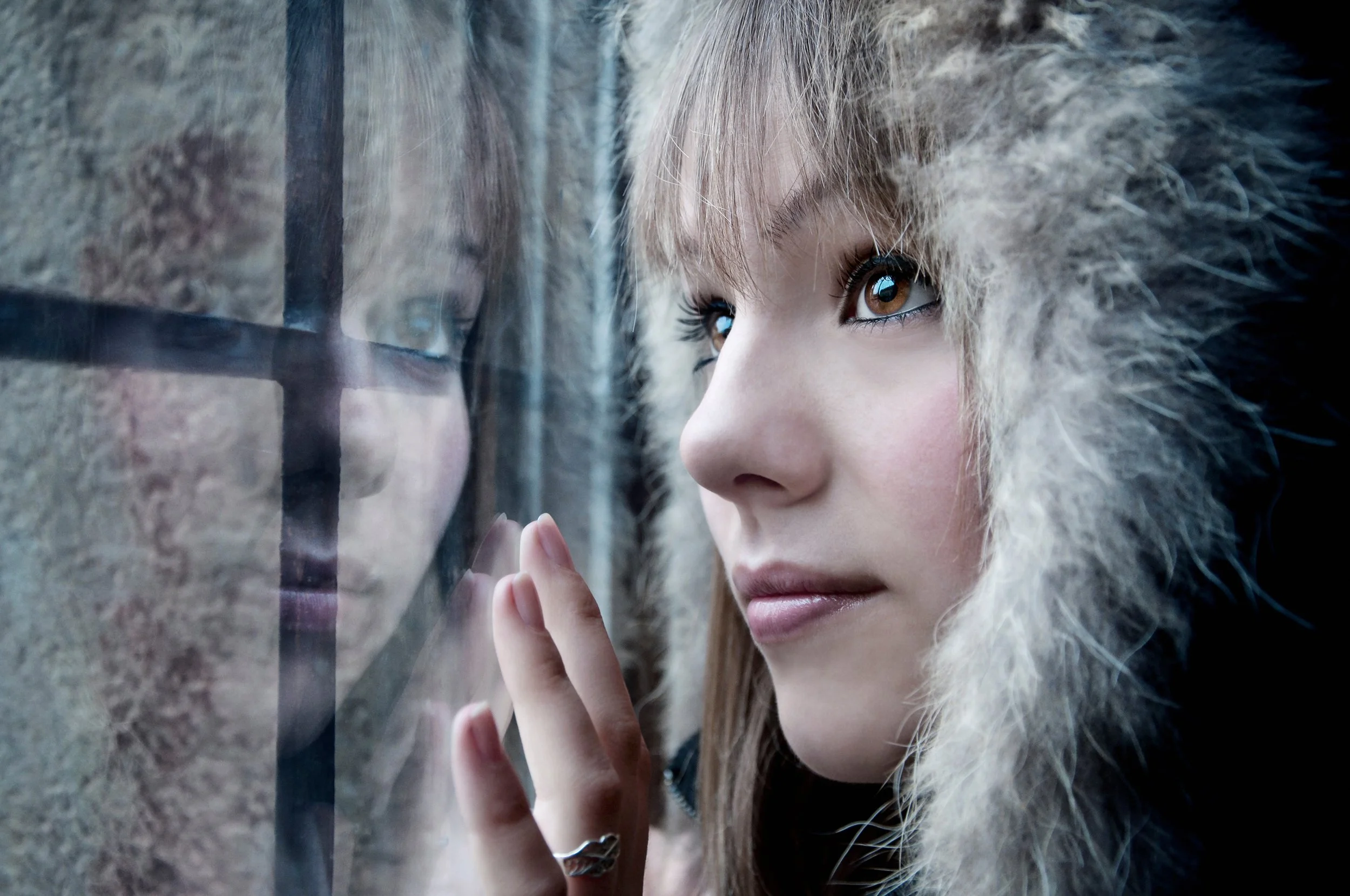 A young woman with brown eyes and light skin looking through a window, wearing a furry hooded coat.