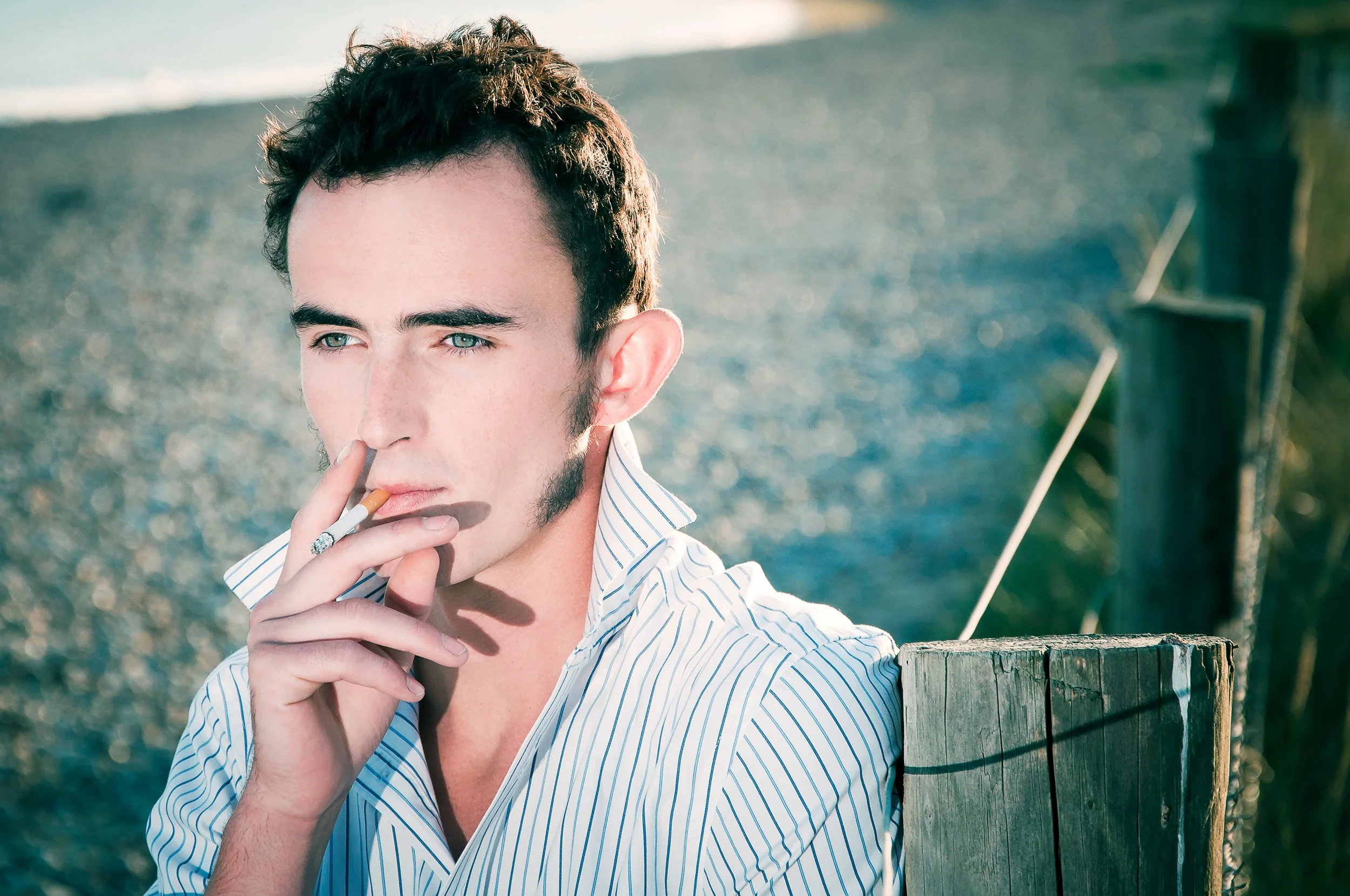 A young man with dark, curly hair and blue eyes is holding a cigarette near his mouth while standing outdoors near a wooden fence, with water and a grassy area in the background.