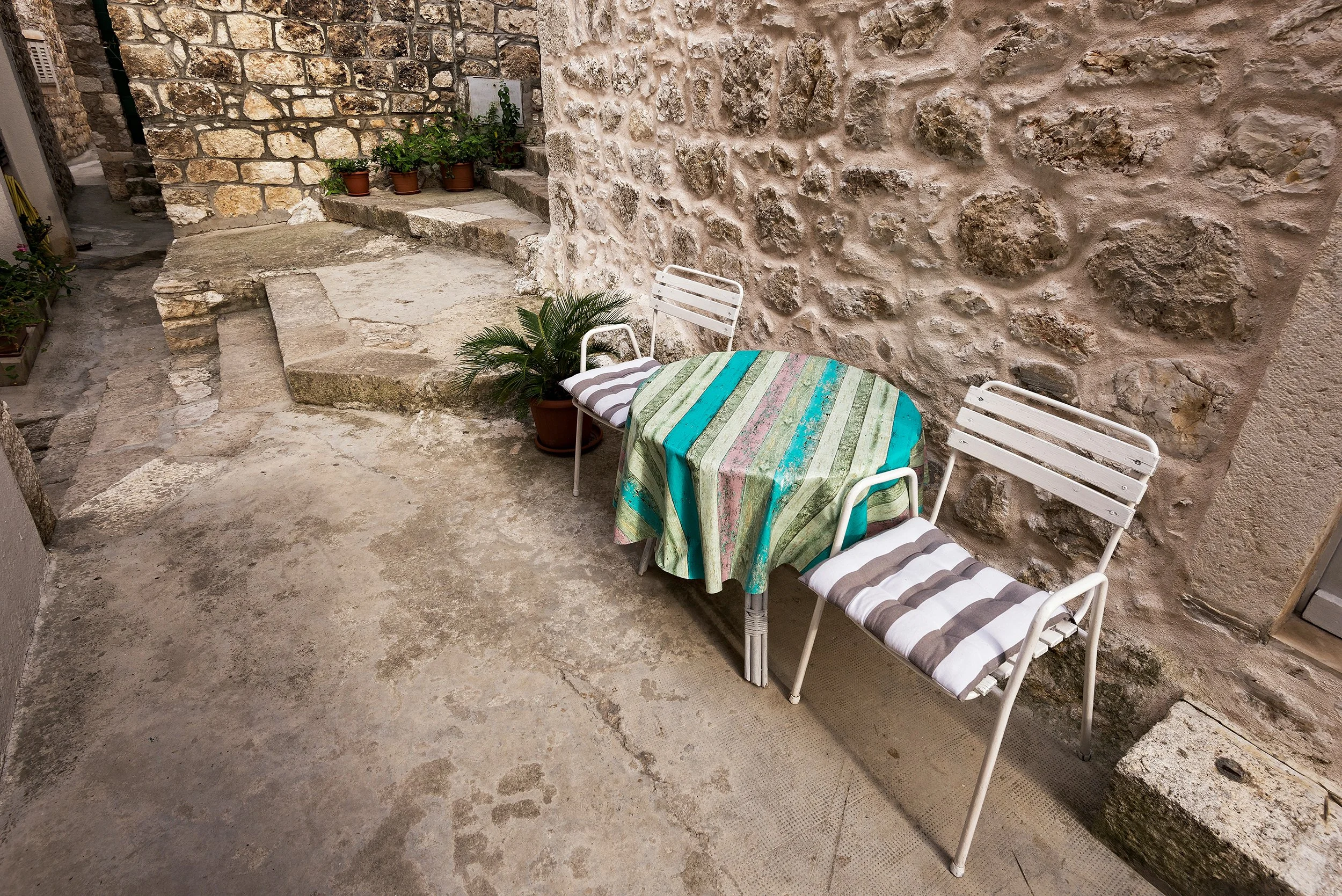 Peter Nemeth photographer. An outdoor patio area with a stone and brick wall, two white metal chairs with striped cushions, a small round colorful table with a striped tablecloth, and several potted plants.