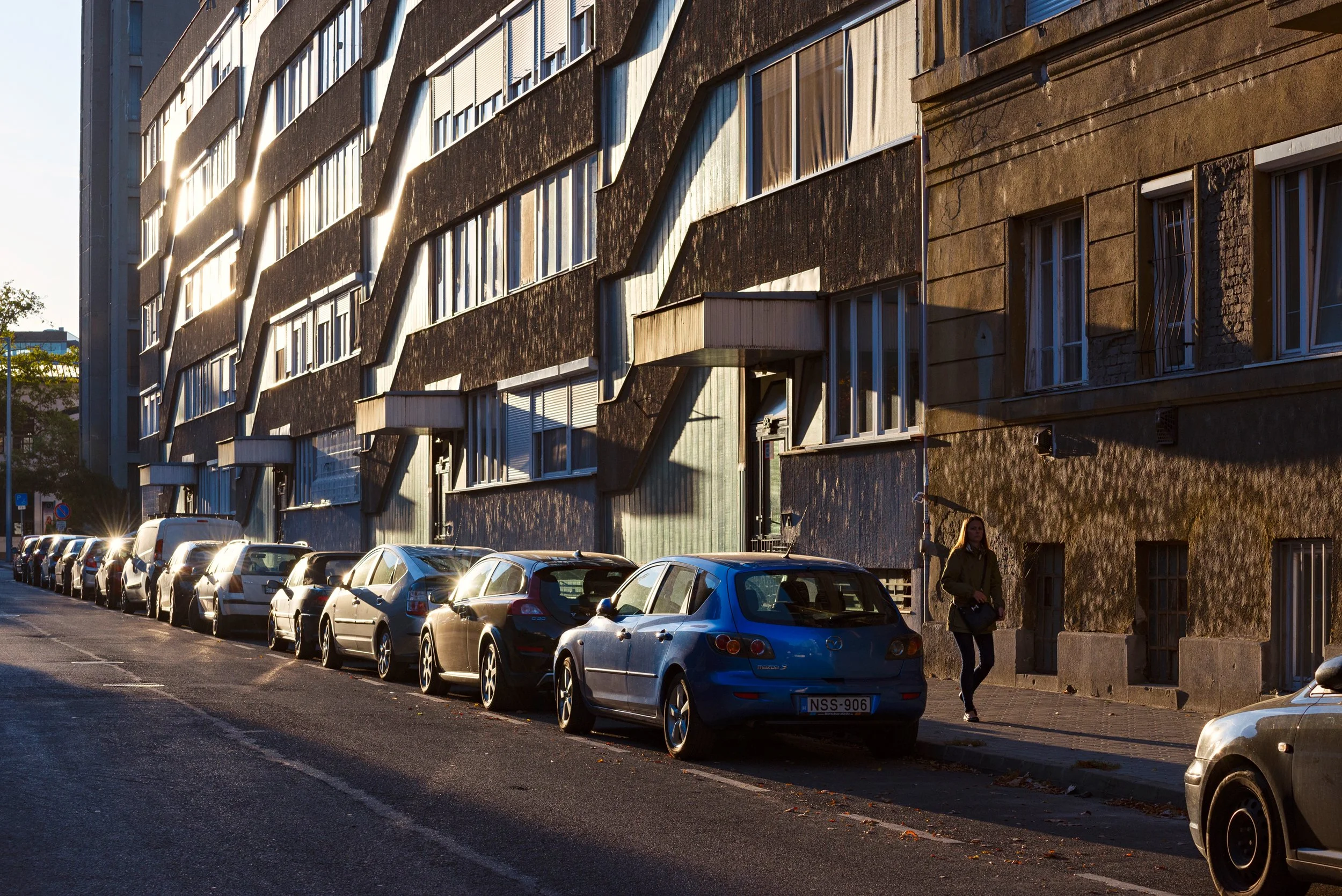 A row of parked cars along a city street with a multi-story residential building in the background. The building has a distinctive architectural design with protruding window frames and a weathered facade, illuminated by the setting sun.