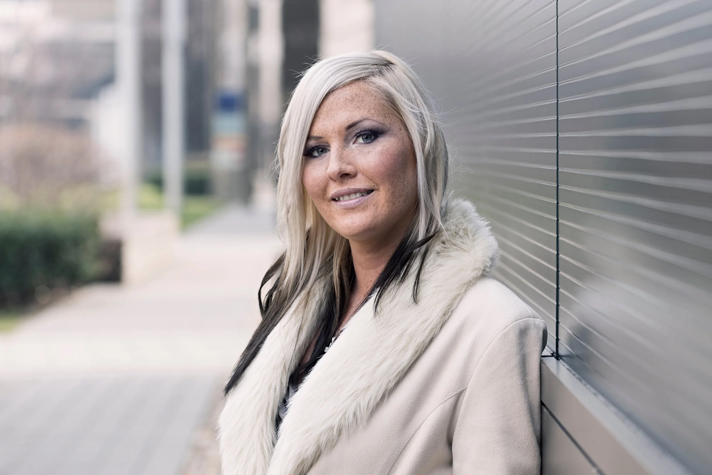A woman with long blonde hair and blue eyes smiling, leaning against a metal wall outdoors, wearing a white fur coat.