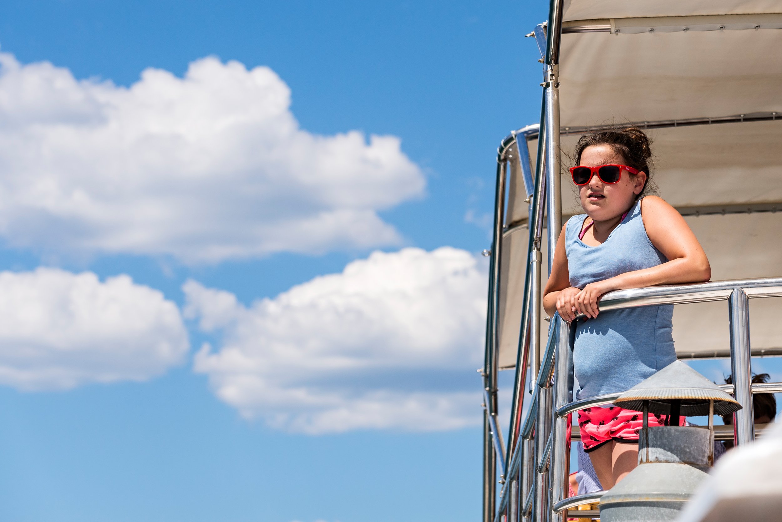A young girl wearing red sunglasses and a blue sleeveless top leaning on the railing of a boat with a clear blue sky and white clouds in the background.