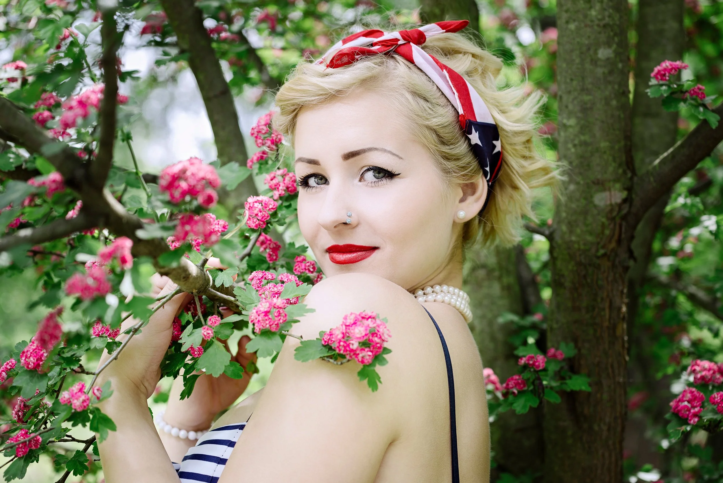 A blonde woman with makeup, red lipstick, pearl earrings, and a pearl necklace, wearing a patriotic red, white, and blue bandana, poses outdoors among pink flowering bushes and green foliage.