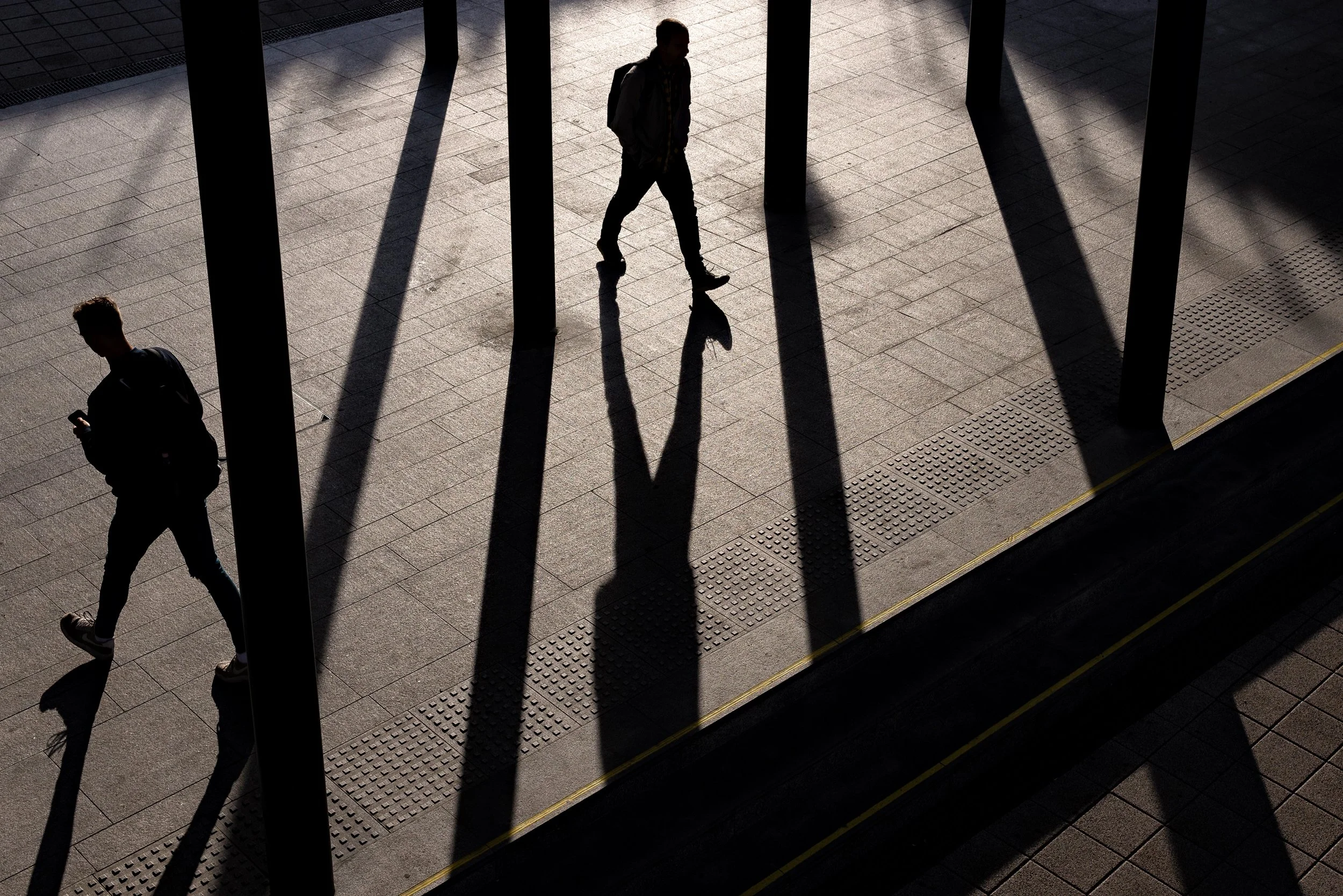 Silhouettes of three people walking on a tiled surface with tall dark shadows cast by vertical beams or structures, sunlight creating high contrast and shadow patterns.