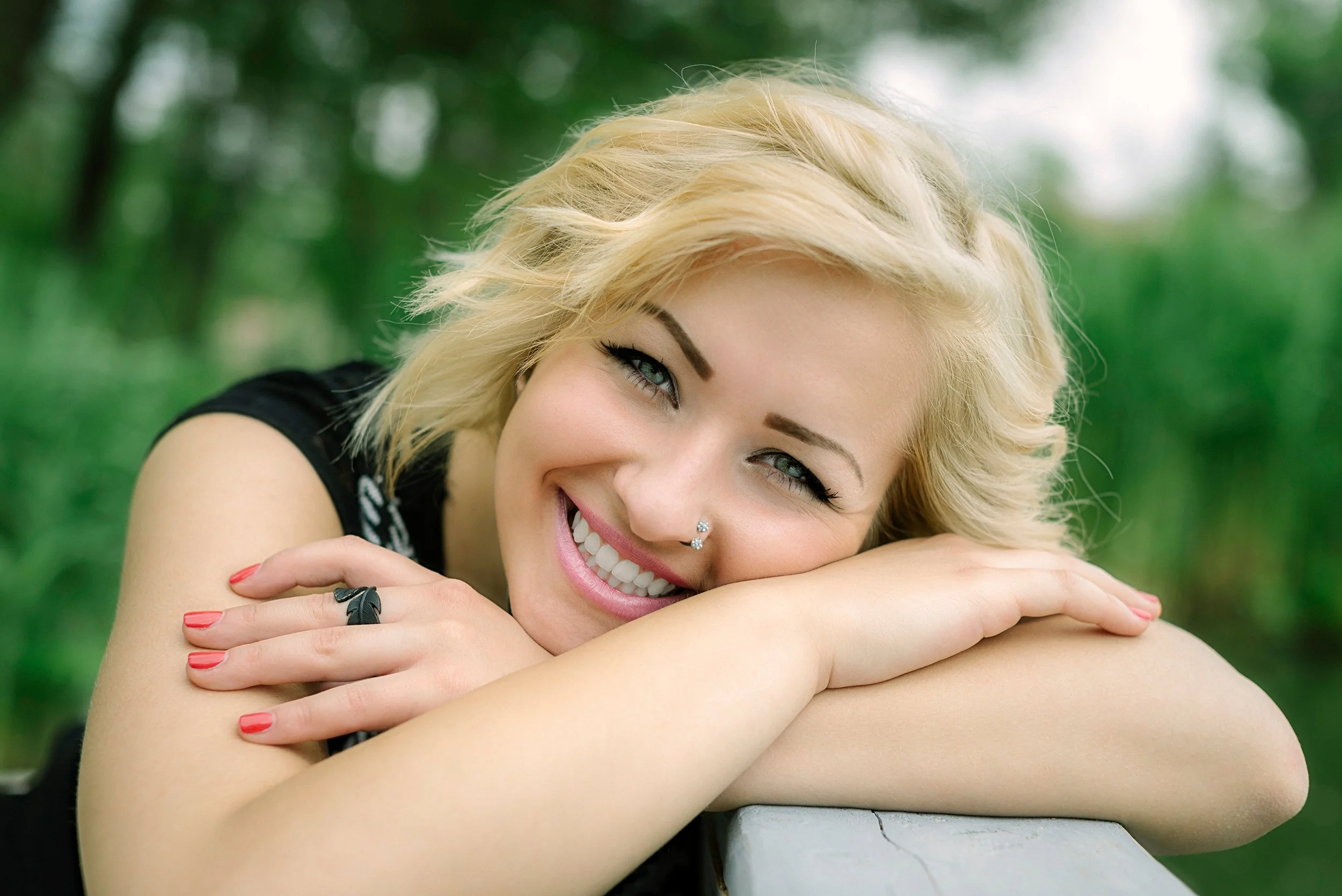 A smiling woman with short blonde hair and a nose piercing, resting her arms on a white surface in an outdoor setting with blurred green trees in the background.