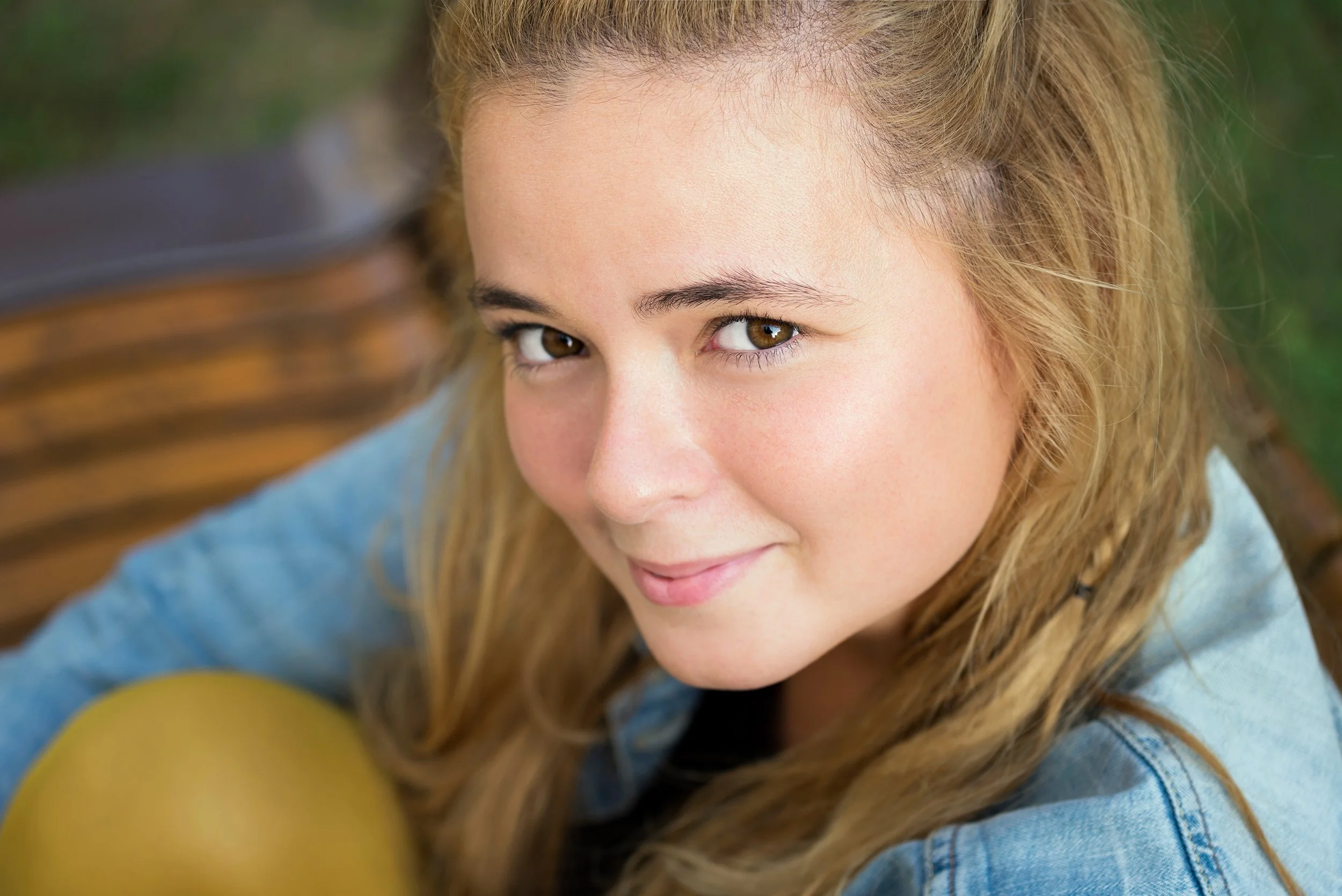 Close-up of a young woman with light brown hair and brown eyes, smiling softly, outdoors with a wooden bench and green foliage in the background.