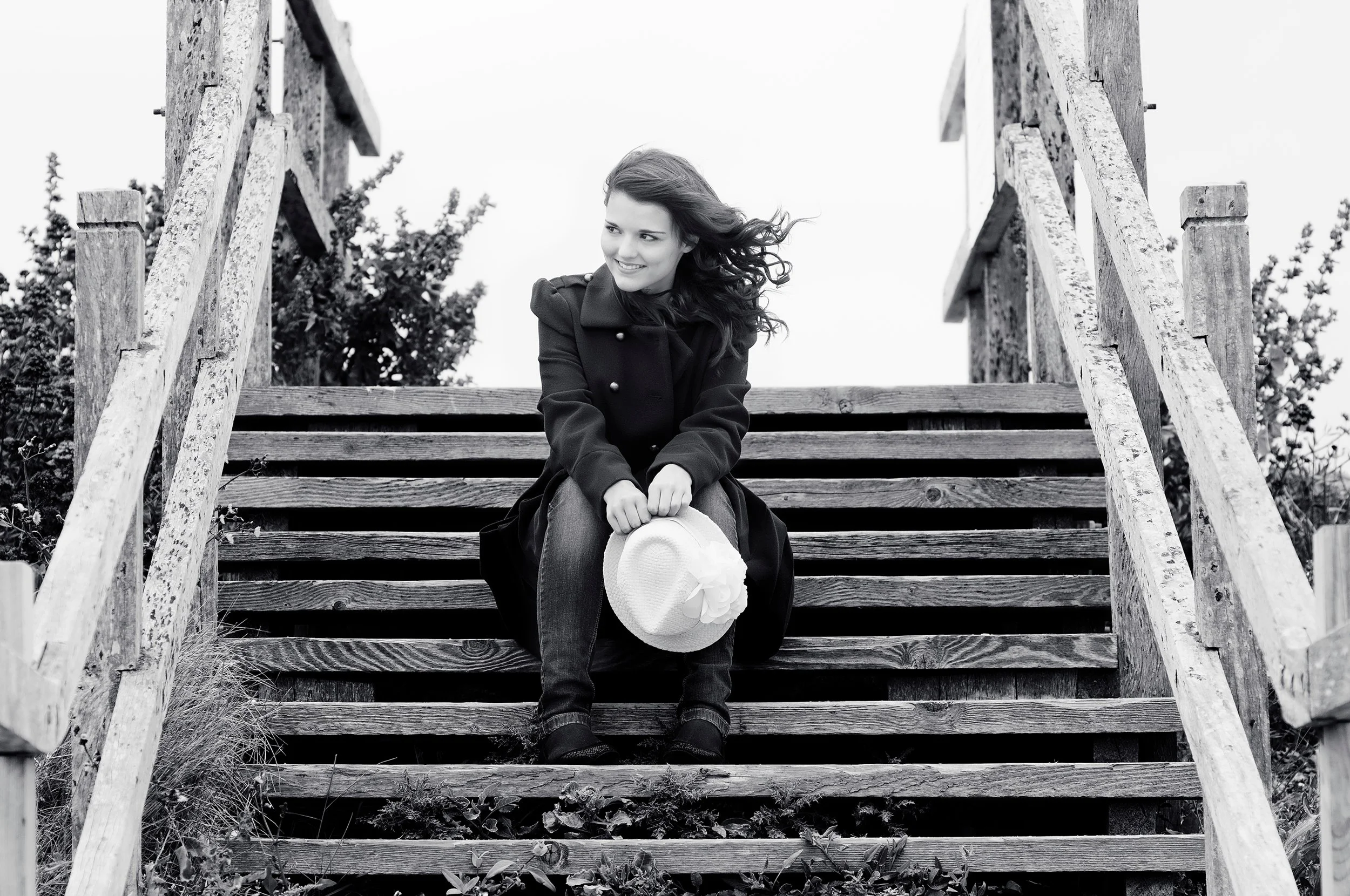 A black and white photo of a young woman sitting on outdoor wooden stairs, holding a wide-brimmed hat, smiling, with her hair blown by the wind.
