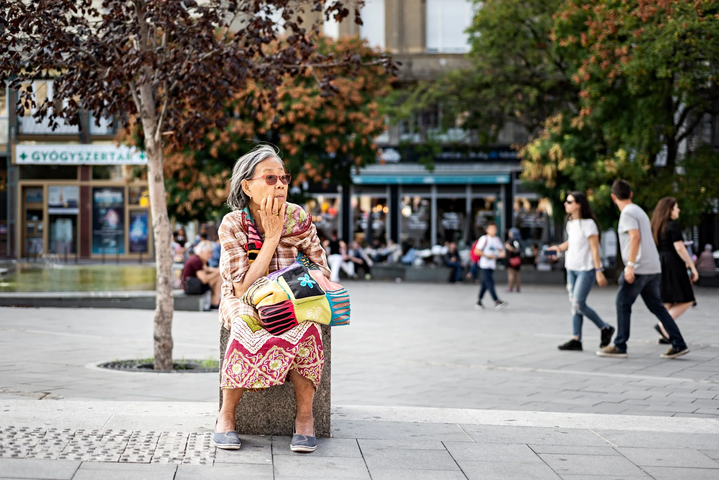 An elderly woman with gray hair, wearing glasses, a colorful patchwork scarf, and a patterned skirt, sits on a stone bench with her hand on her chin in an urban park. There are trees with autumn-colored leaves and a busy city background with pedestri