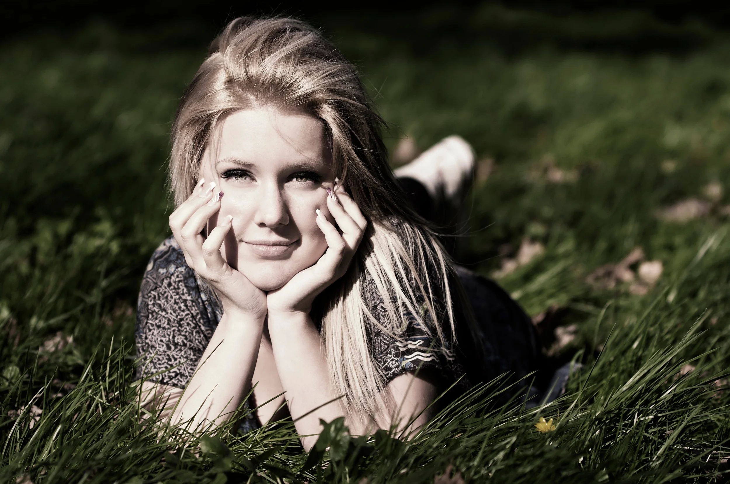 A young woman with long blonde hair lying on grass outdoors, resting her chin on her hands and looking at the camera with a slight smile.