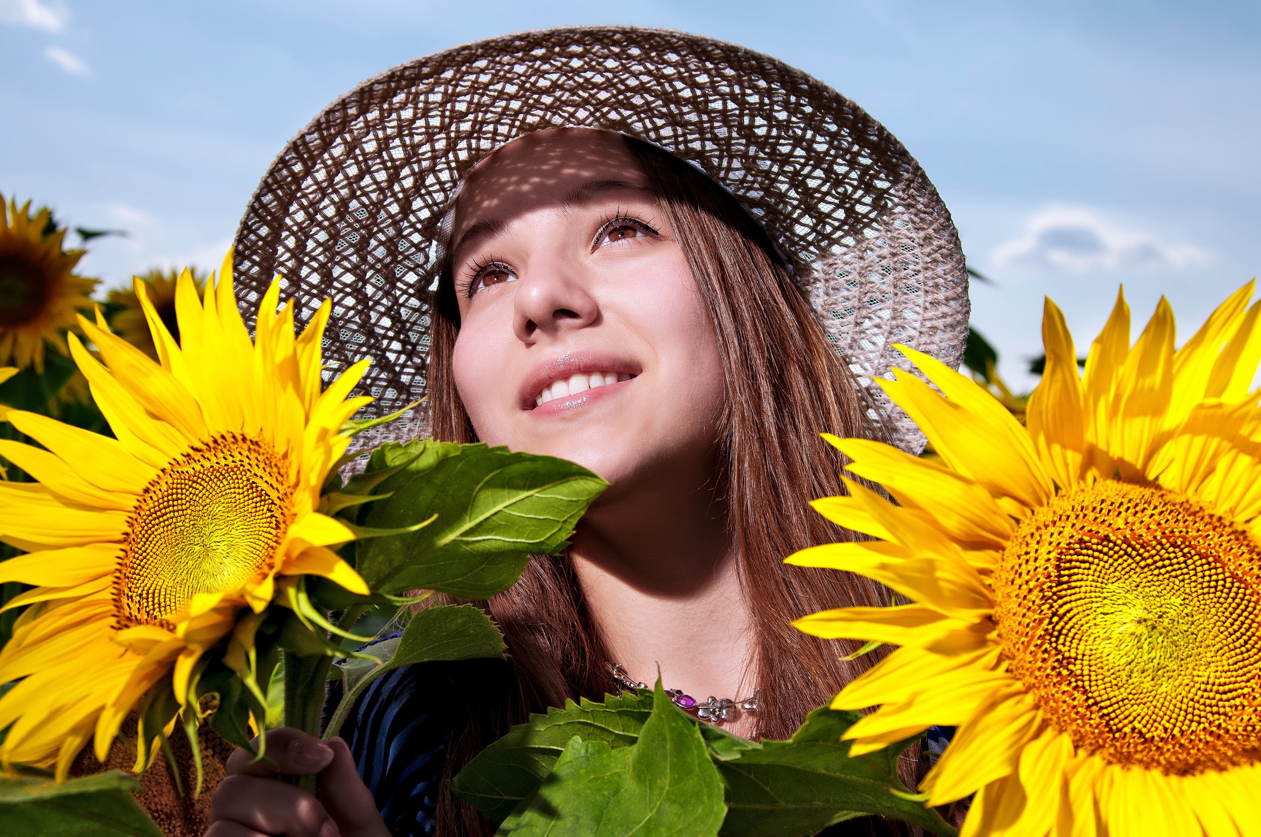 A young woman with long brown hair and light skin smiling, wearing a sun hat and jewelry, holding sunflowers in a bright outdoor setting with blue sky and clouds.