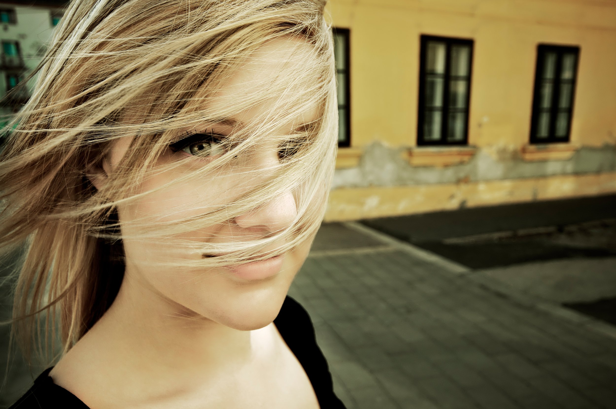 Close-up of a woman with blonde hair partially covering her face, standing outdoors with a building featuring several windows in the background.