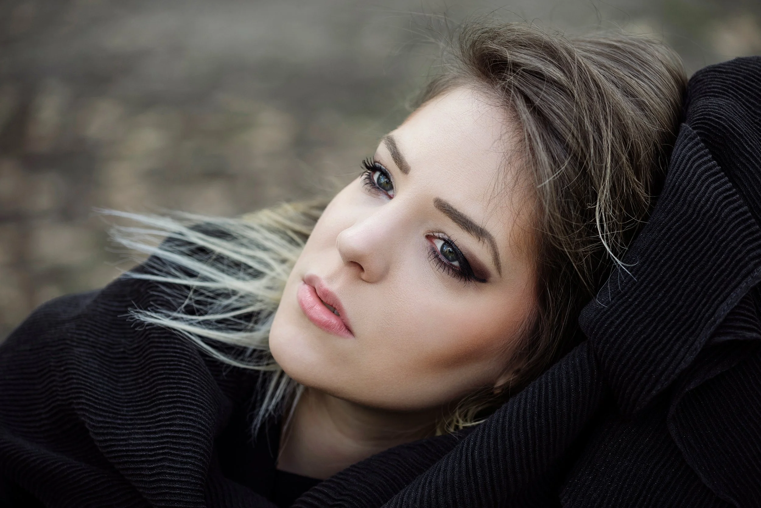 A woman with makeup and light-colored hair lying down outdoors, with her head tilted and eyes gazing forward.